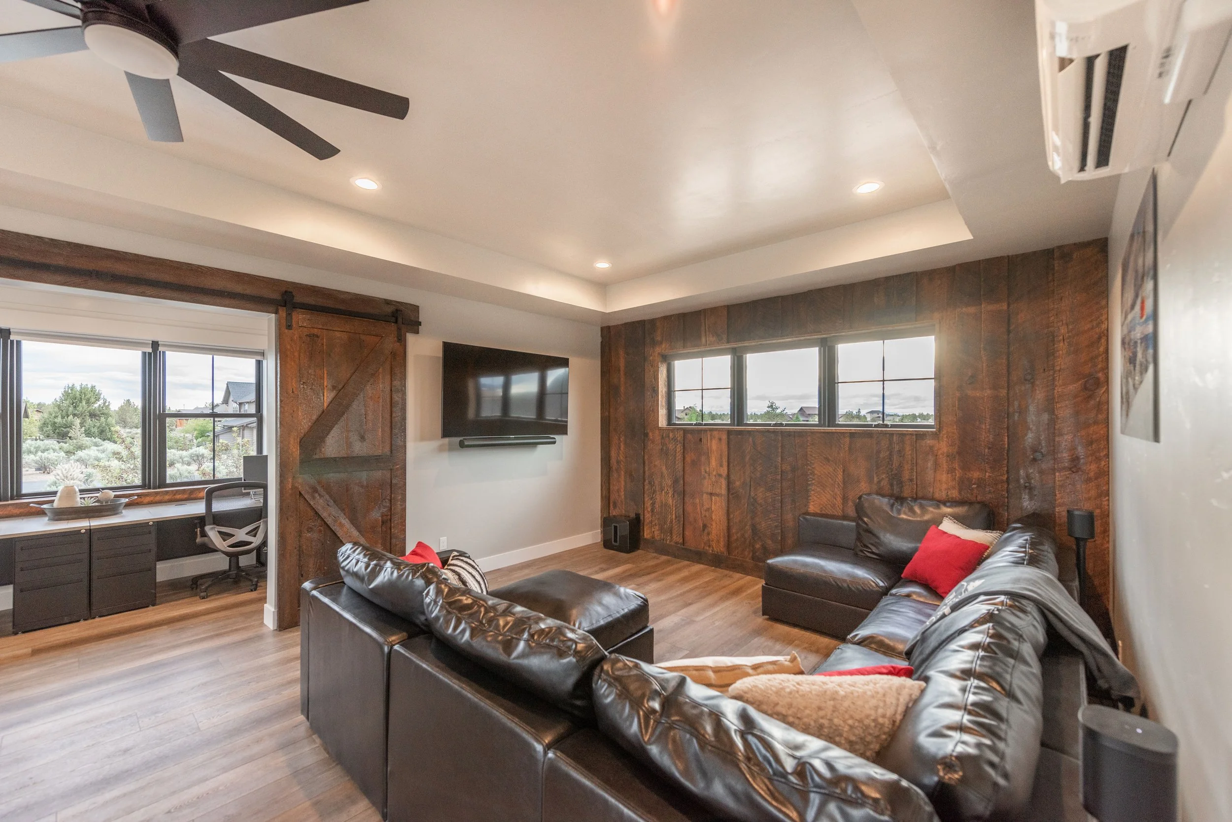 Living room with brown leather sectional sofa, wooden wall paneling, and a window view of outdoors. A flat-screen TV is mounted on a light-colored wall. There is a ceiling fan, recessed lighting, and a corner desk with an office chair near the window