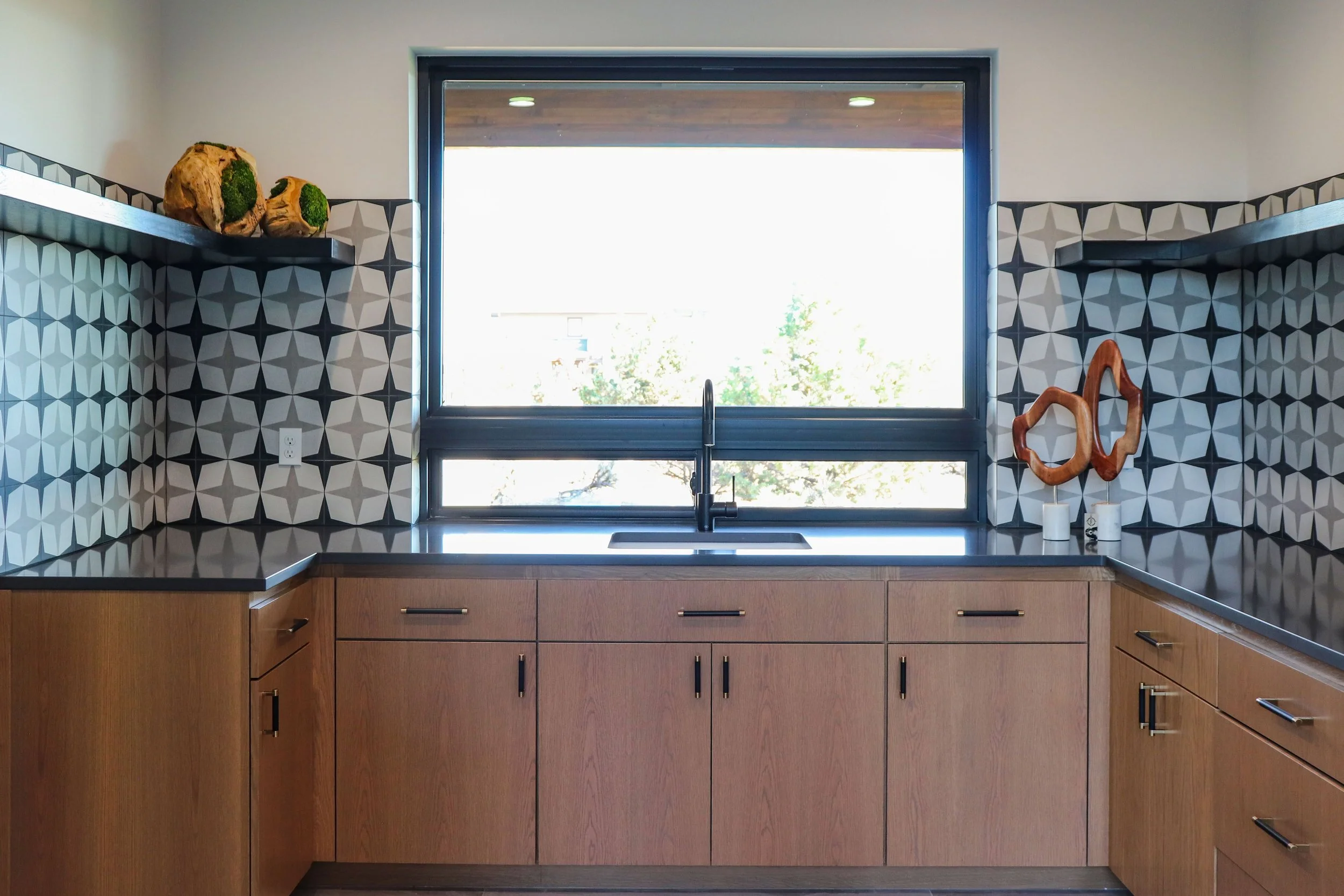 Modern kitchen with wooden cabinets, black countertop, geometric black and white tile backsplash, large window above the sink, decorative wood sculptures, and potted plants on shelves.