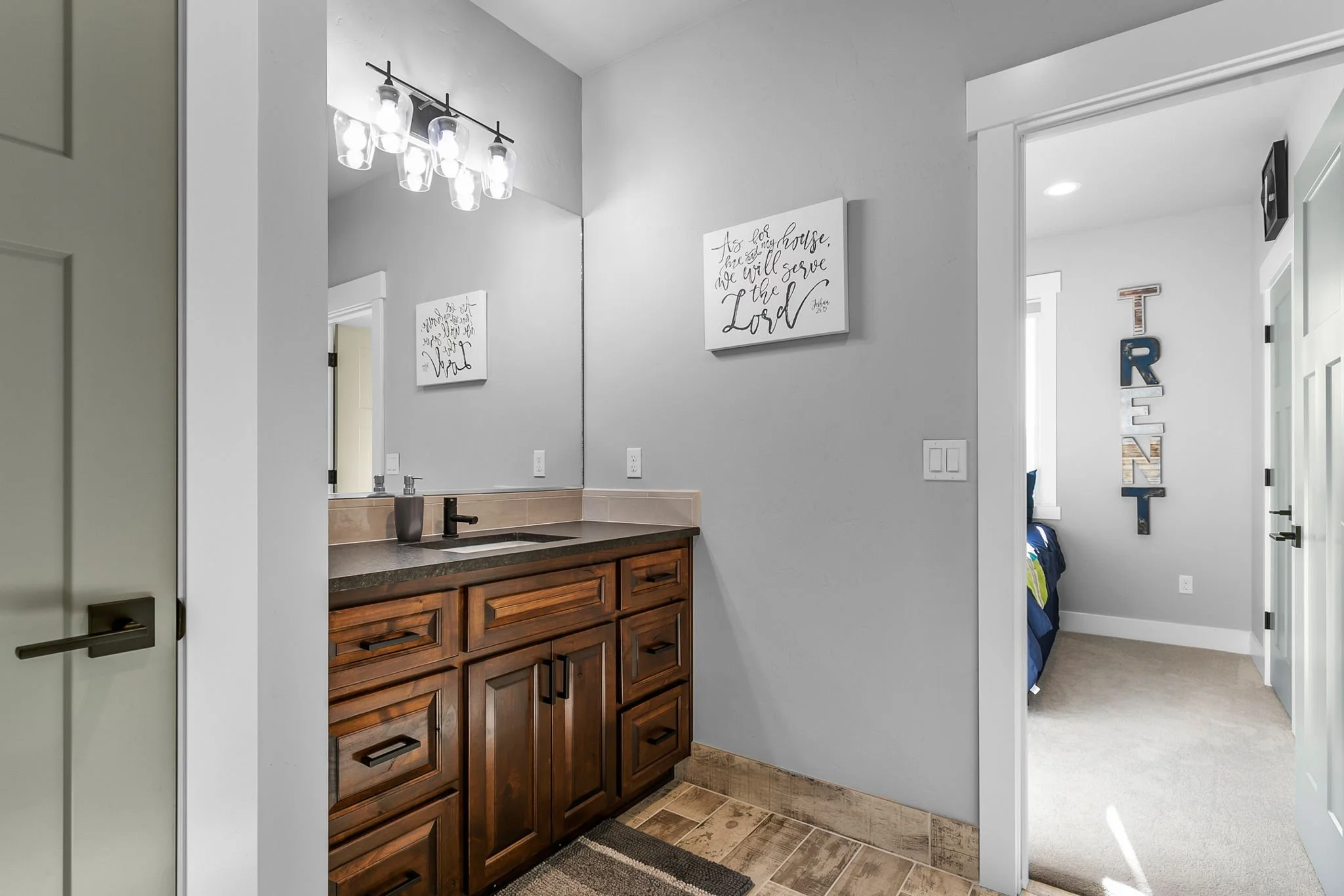 Bathroom vanity with wooden cabinet, gray countertop, and a large mirror. Light fixture with multiple bulbs above the mirror. Wall art with quote on the wall to the right of the mirror. Door to the right leads to a bedroom with wall decor spelling 'T