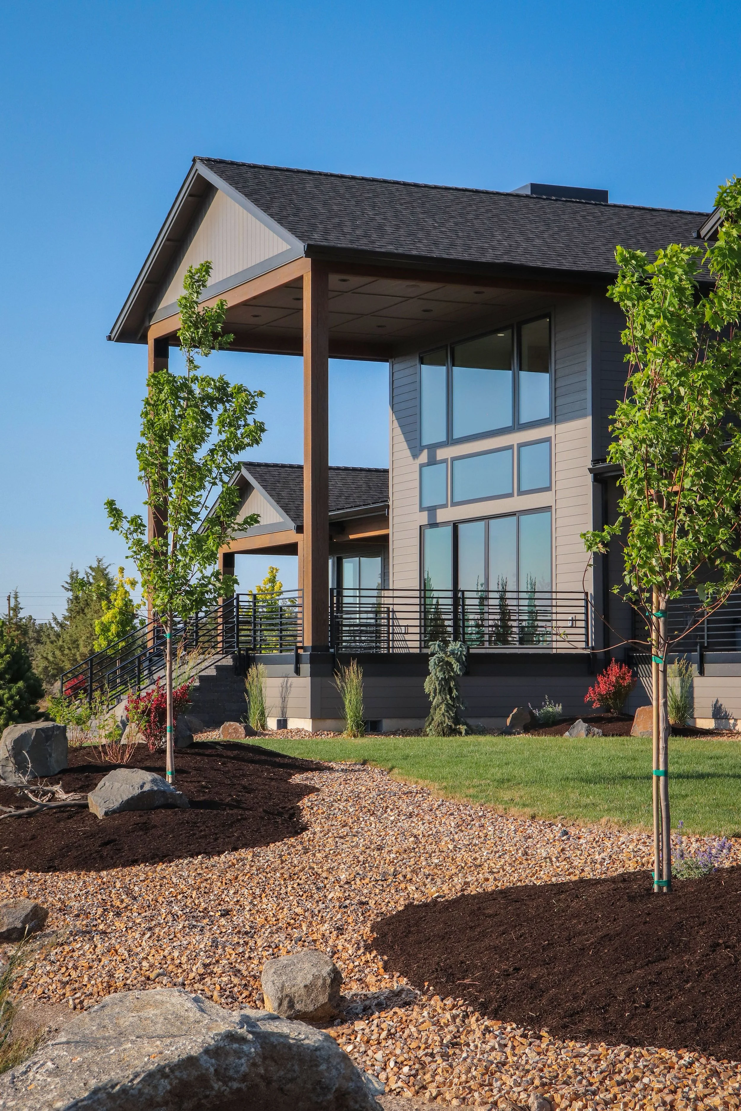 Modern house with large windows, surrounded by landscaped yard with young trees and rocks, under a clear blue sky.