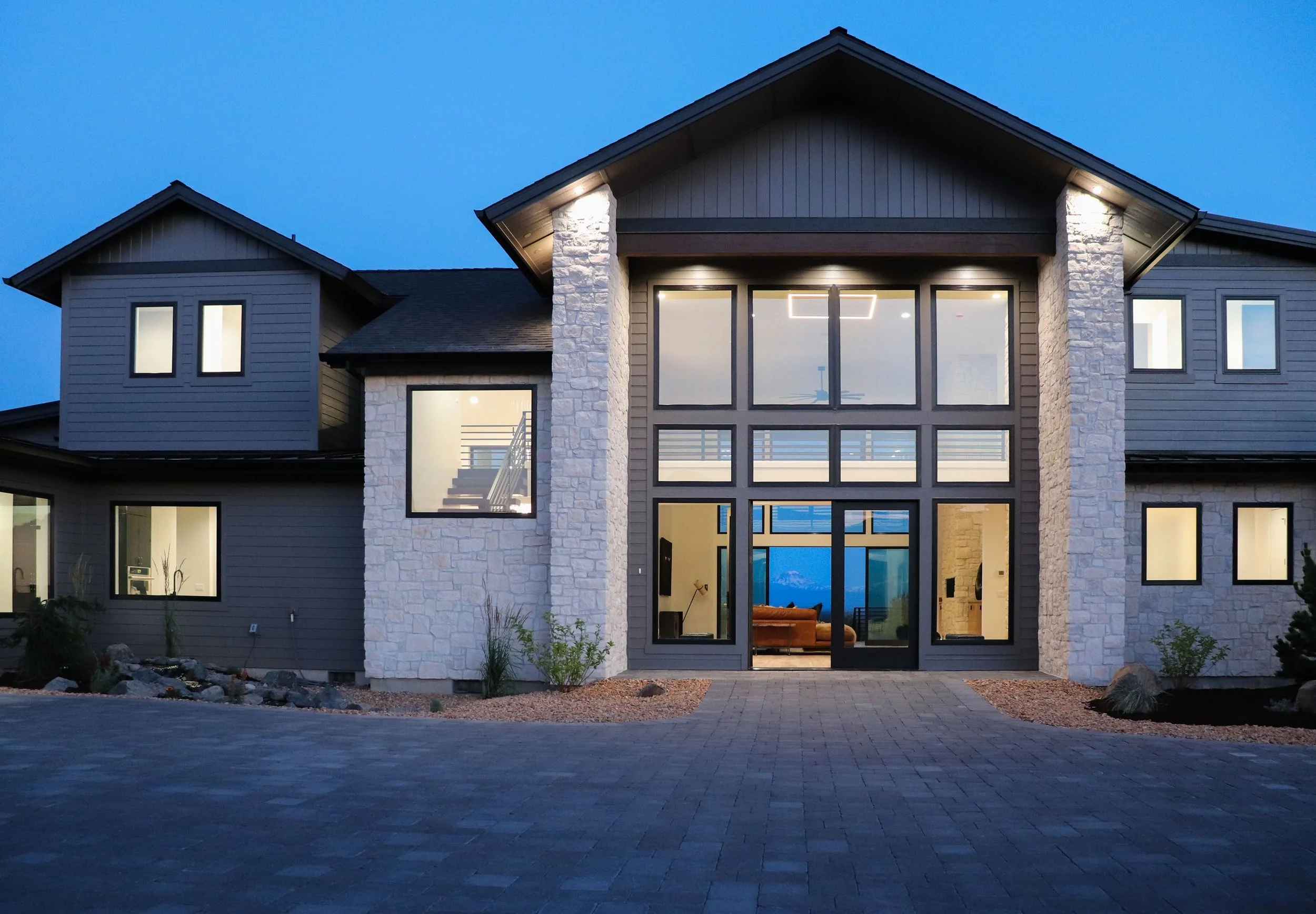Modern two-story house with large glass windows, stone and siding exterior, and illuminated interior, at dusk.