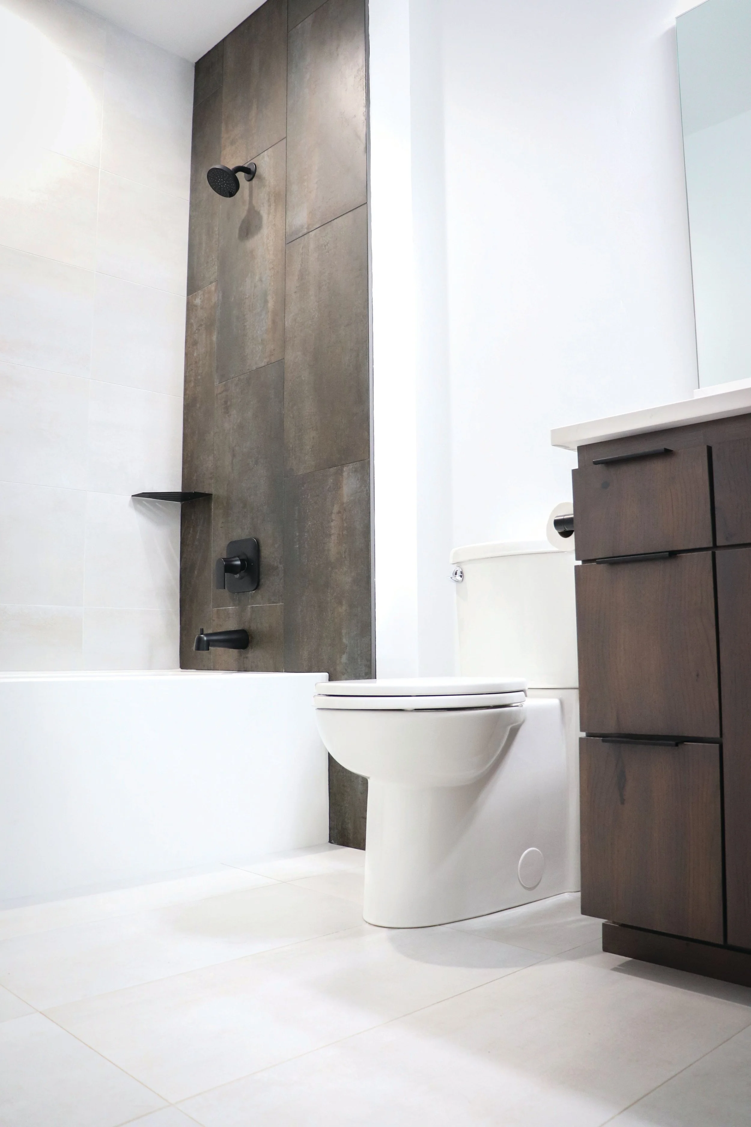 Modern bathroom with a white toilet, a dark wood vanity with black handles, and a tile shower area with dark gray tiles and a black showerhead.