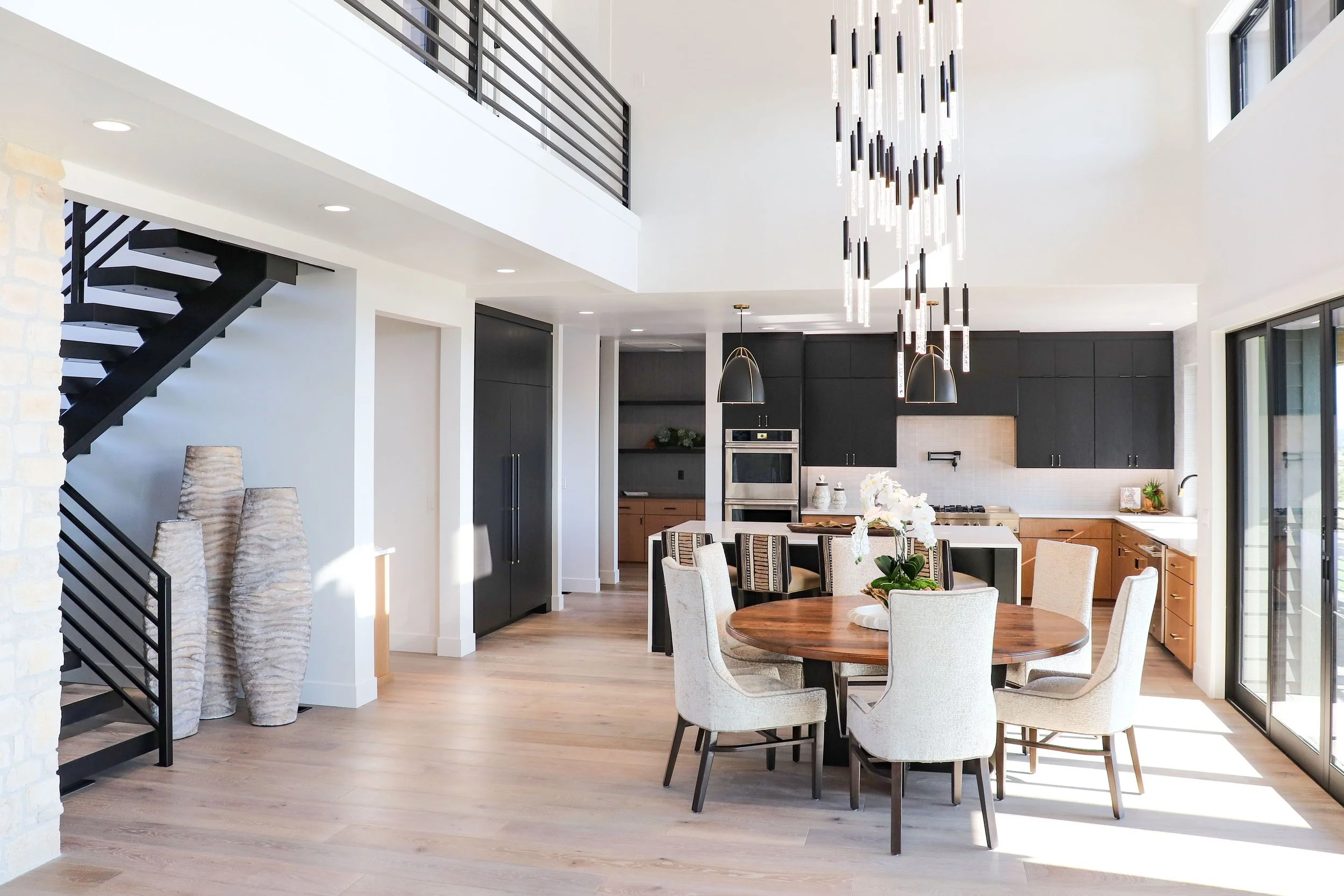 Modern open-concept kitchen and dining area with black cabinets, stainless steel appliances, a round wooden dining table with six chairs, and a large hanging light fixture, with sliding glass doors providing natural light.