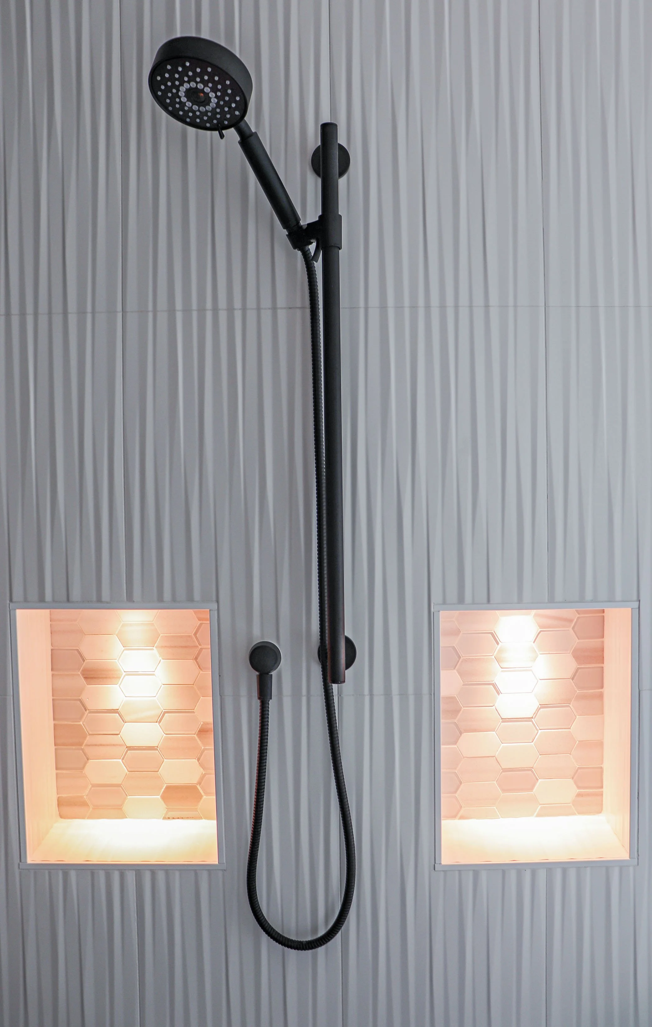 Modern black showerhead with handheld shower on a gray textured wall with two built-in shelves illuminated by warm lighting.