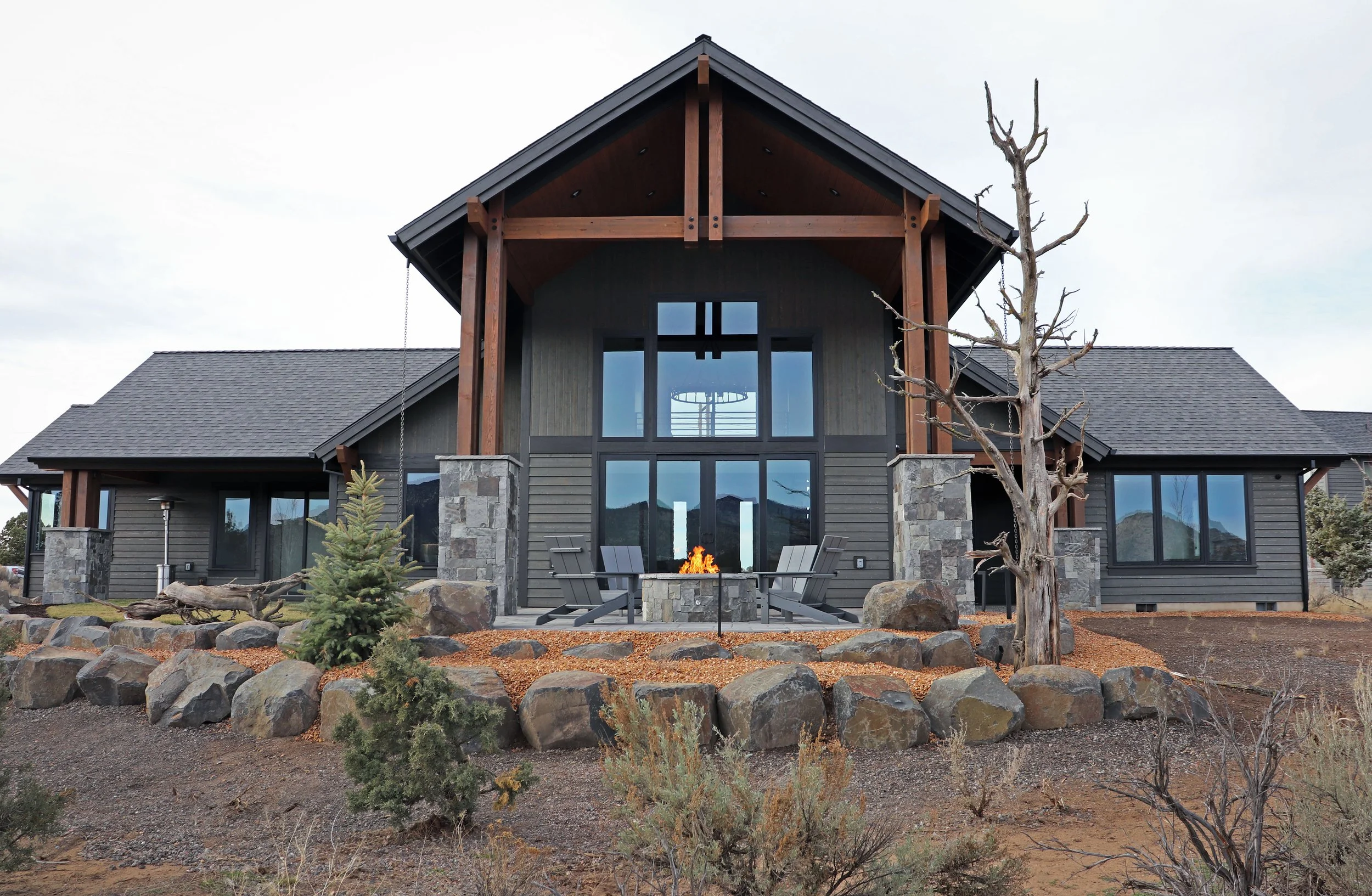 Modern house with large windows, wooden beams, gray siding, stone foundation, and an outdoor fire pit with chairs. There are small trees and rocks in front, and the sky is overcast.