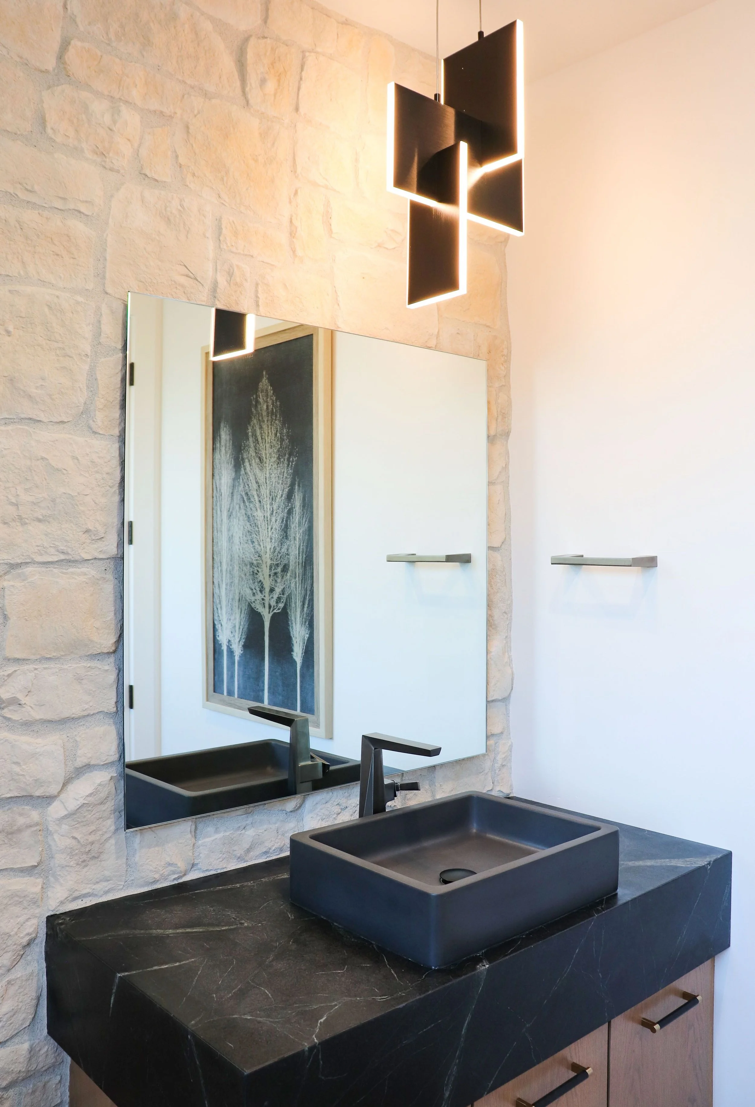 Modern bathroom with a black square vessel sink, black faucet, large mirror, stone wall, and contemporary black ceiling light fixture.