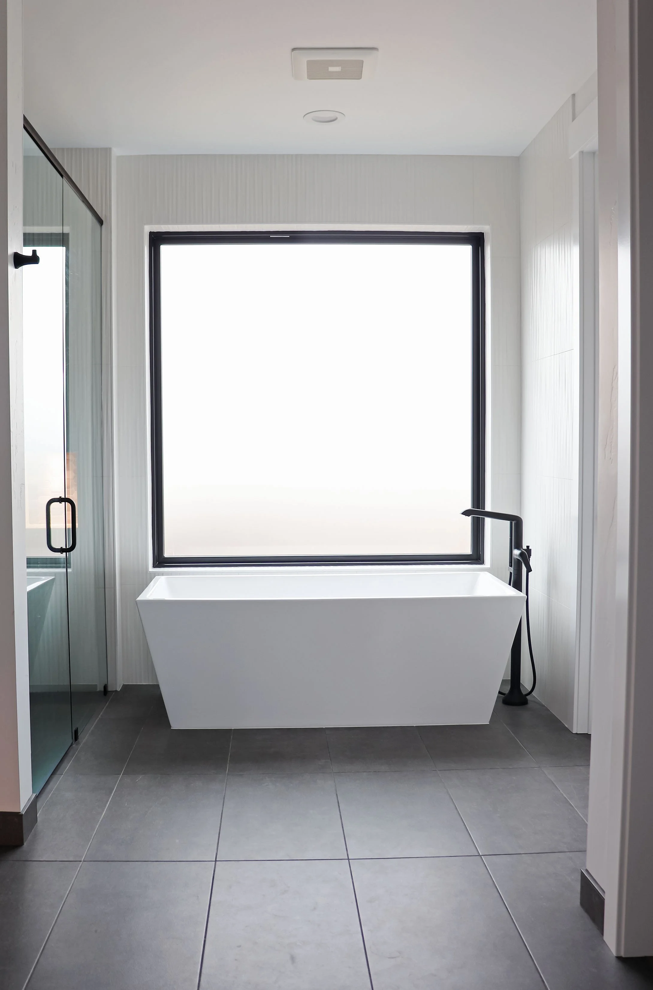 Modern bathroom with a white bathtub, large window, black floor faucet, gray tiled floor, and corner shower with glass door.