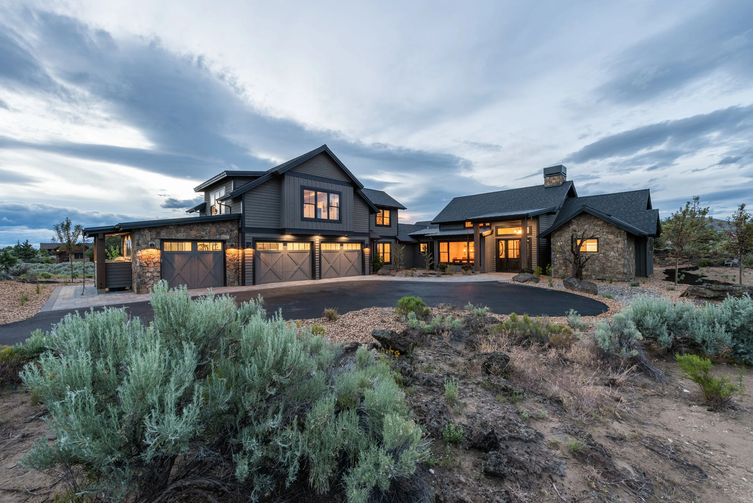 A large modern house with dark exterior and illuminated windows, set on a landscaped yard with desert plants, under a partly cloudy sky at dusk.