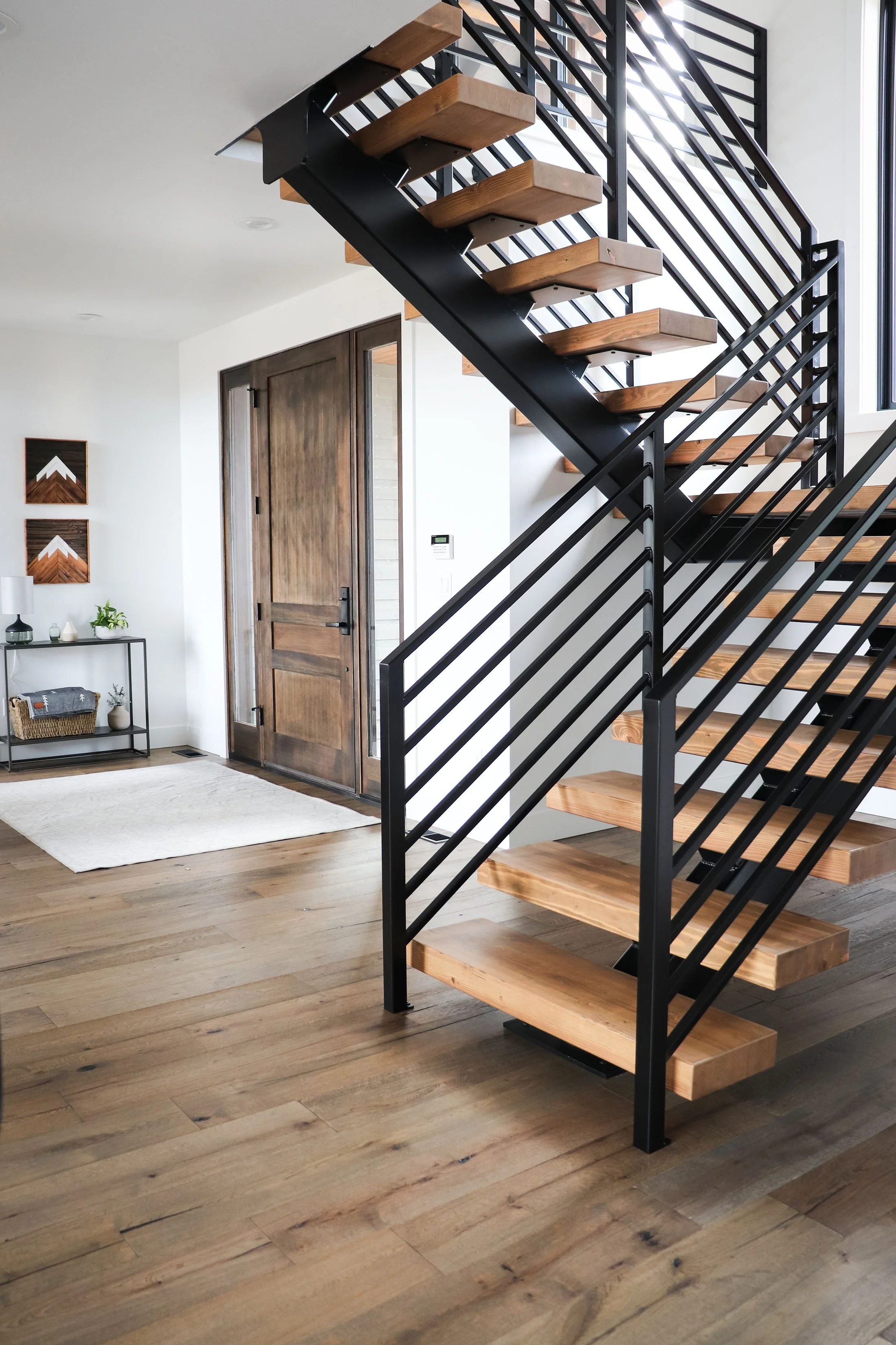 Interior of a modern home with a staircase that has wooden steps and black metal railings, next to a wooden front door and a small entryway table.