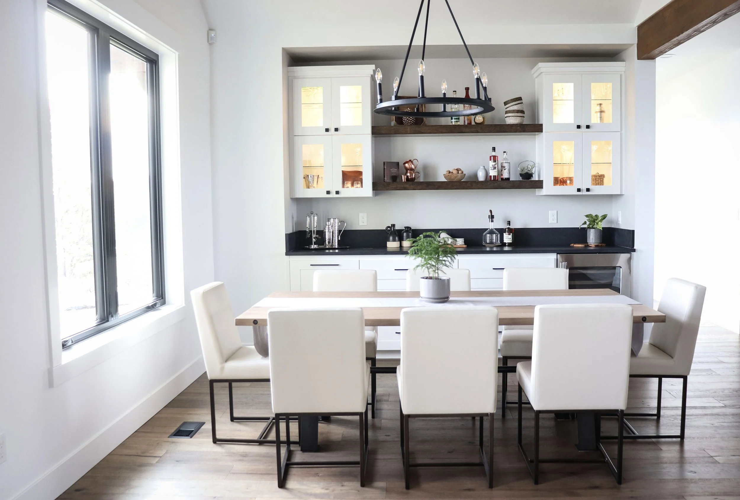 Modern dining area with white chairs around a wooden table, a potted plant in the center, large window on the left, and a kitchen with white cabinets and open shelves in the background.