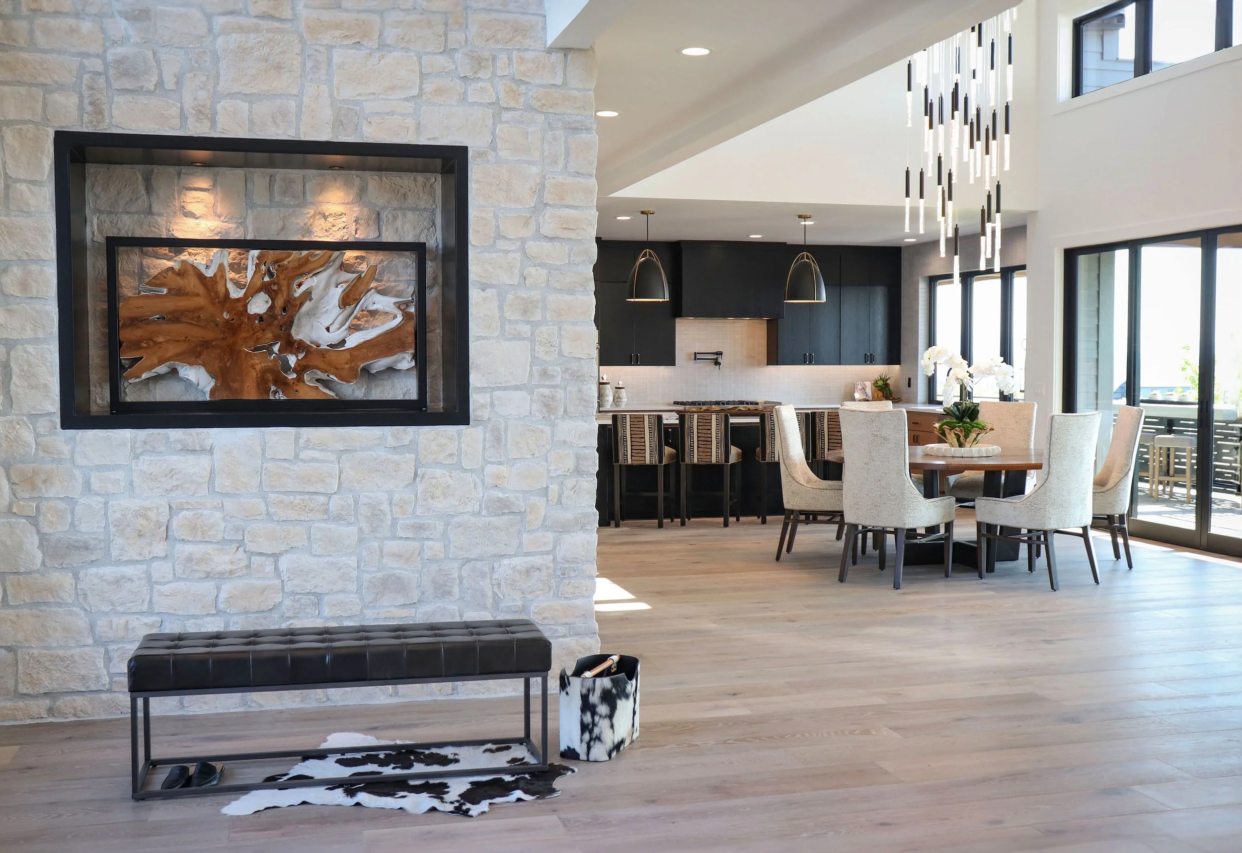Open-concept living area with a stone accent wall, a leather bench, a cowhide rug, and a cat-themed storage bin in the foreground. The dining area has a centerpiece with white flowers, and the kitchen has dark cabinets, a bar with striped stools, and