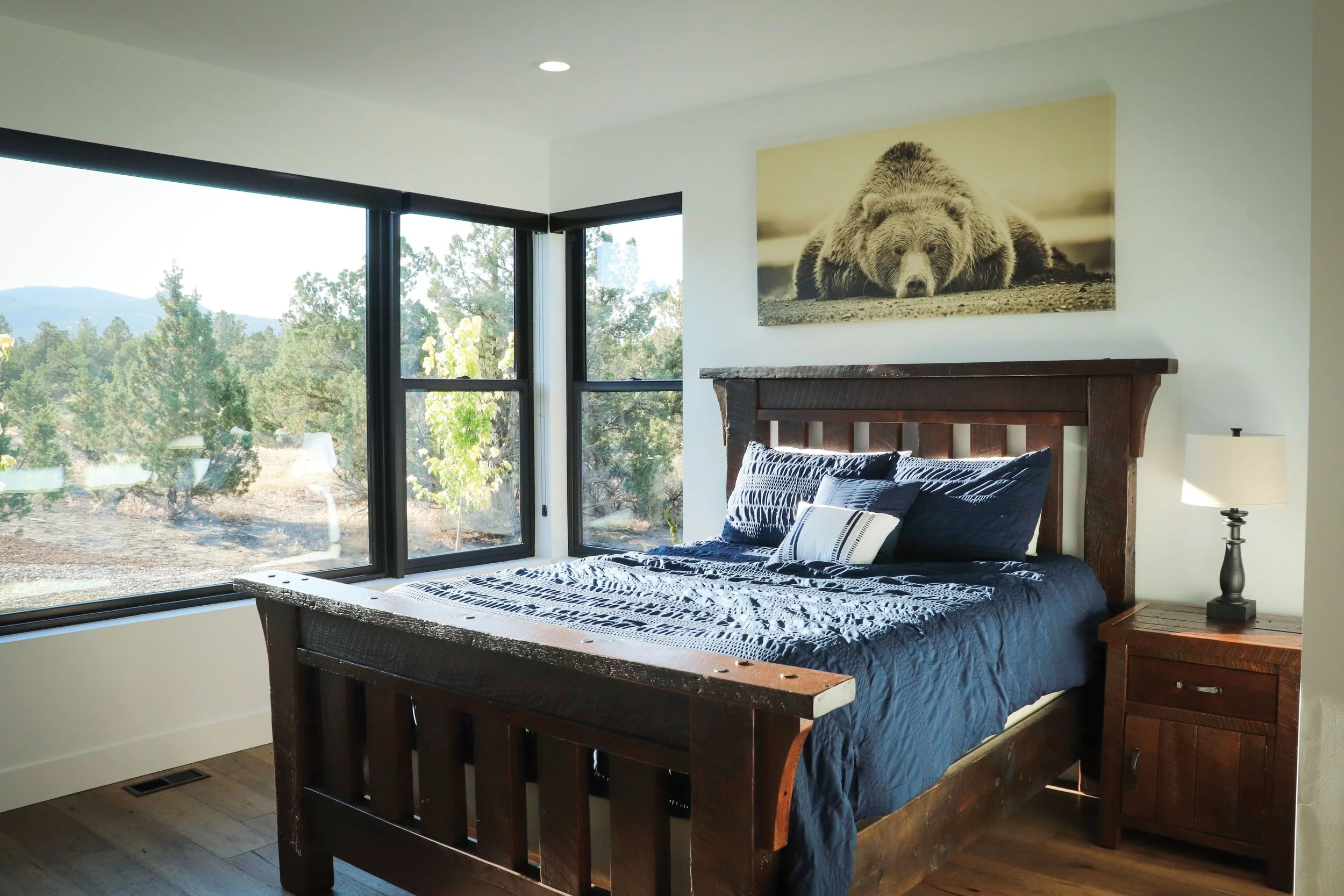 Bedroom with large windows, wooden bed frame with blue bedding, nightstand with lamp, and bear picture on the wall.