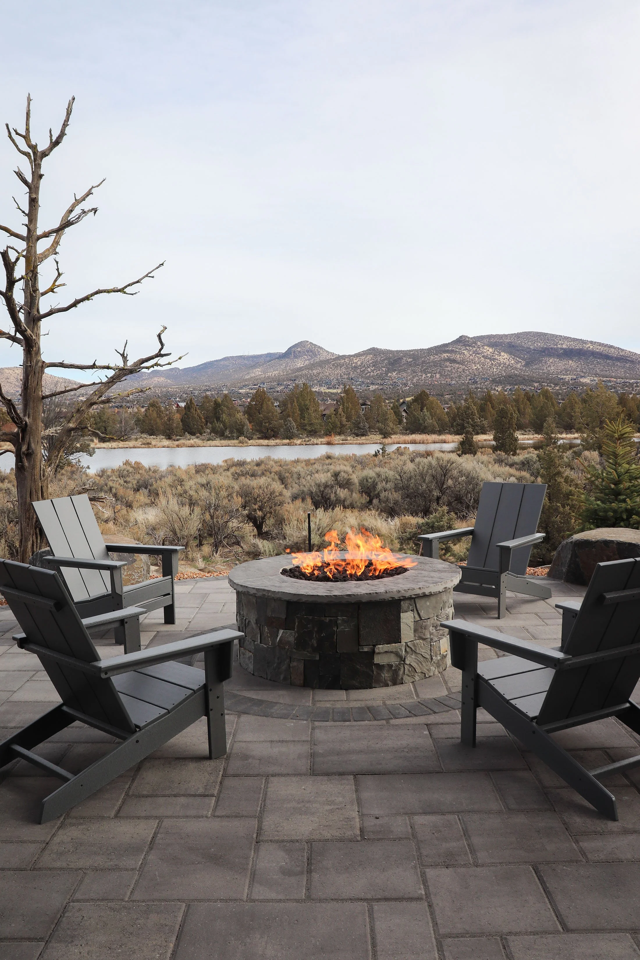 Four gray outdoor chairs arranged in a circle around a stone fire pit on a patio, overlooking a lake, trees, and mountains in the distance.