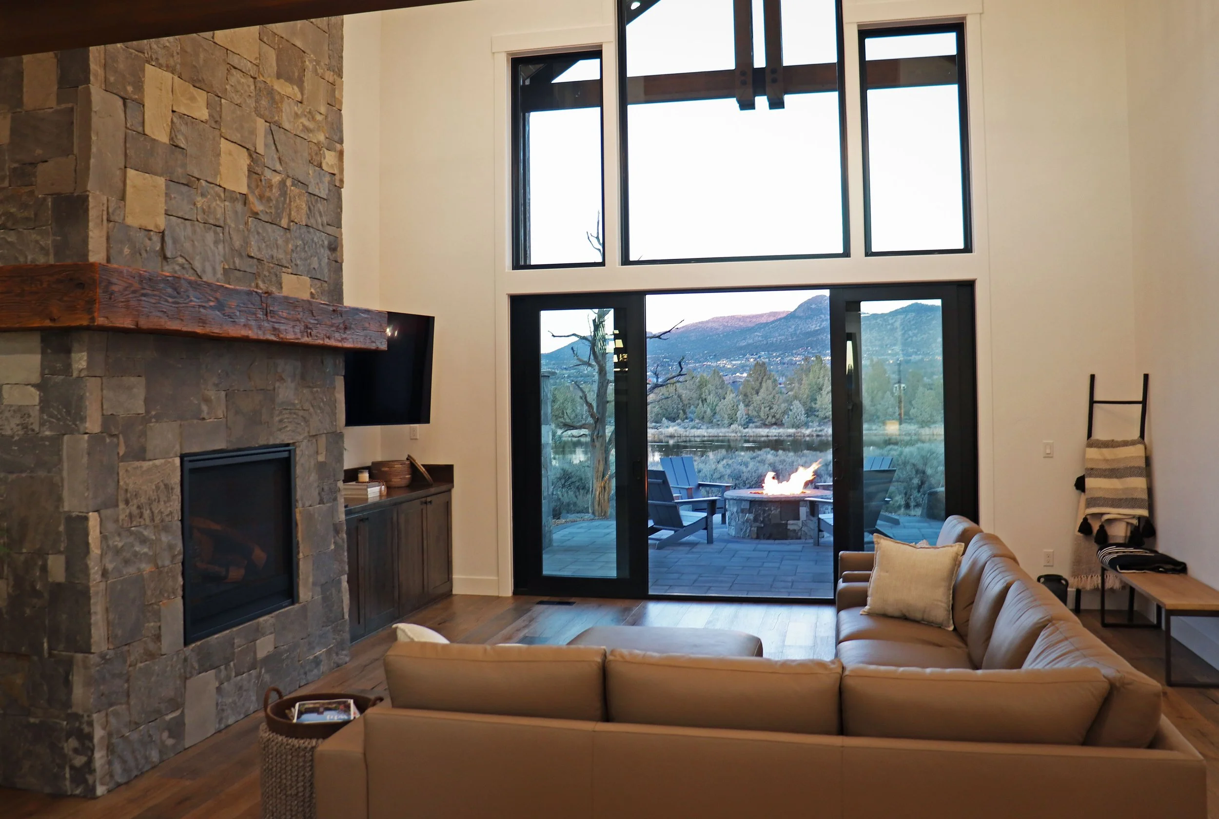 Living room with large windows showing outdoor scenery of trees and mountains, fireplace, beige sectional sofa, and outdoor fire pit with chairs.