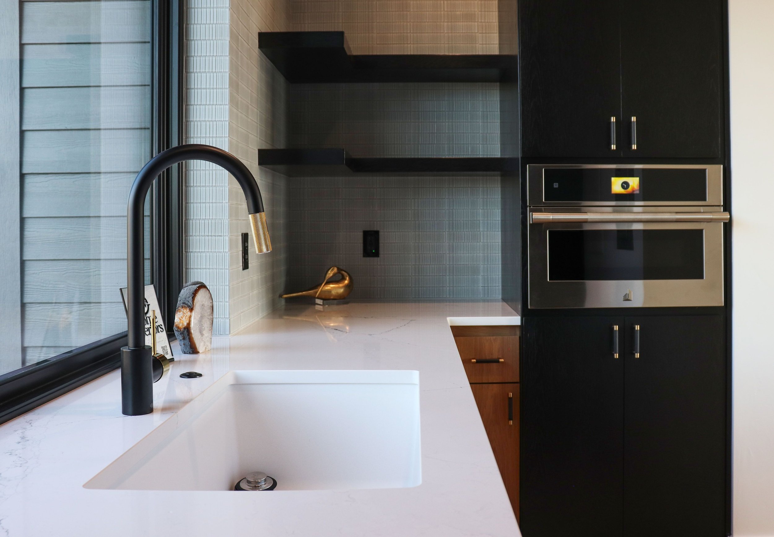 Modern kitchen with white marble countertop, black faucet, window, black shelving, black cabinets, built-in microwave, and decorative gold duck sculpture.