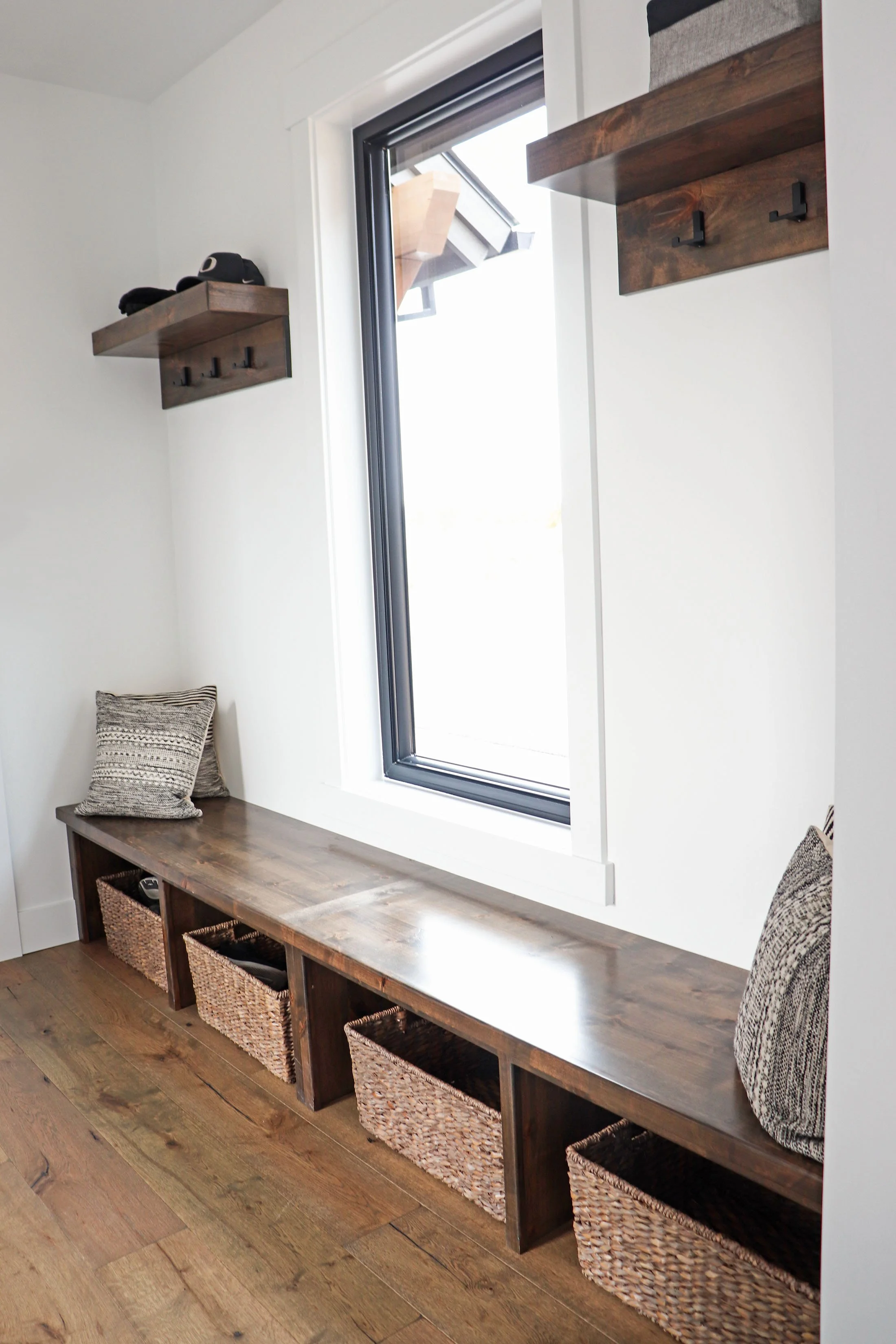 A cozy window seat with built-in storage baskets beneath a large window, decorated with patterned cushions, in a modern interior with white walls and wooden accents.