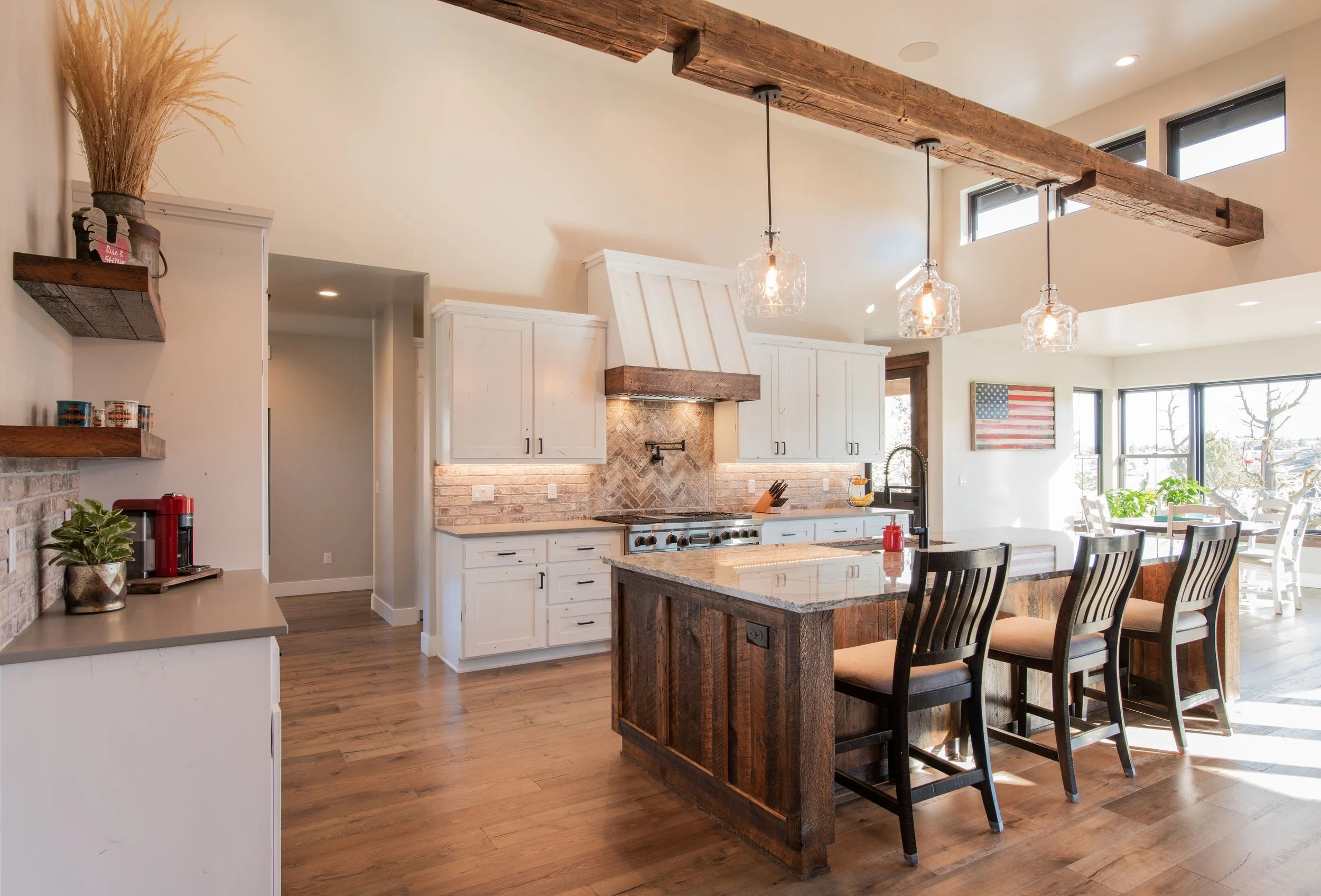 Open-concept kitchen with white cabinetry, a rustic wooden kitchen island, pendant lighting, large windows, and a dining area in the background decorated with an American flag wall art.
