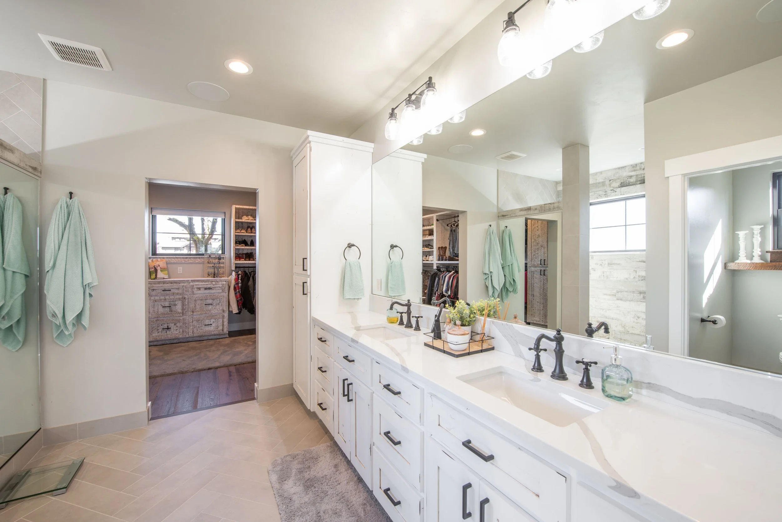 Bright bathroom with large mirror, double sink vanity, white cabinets with black hardware, two soap dispensers, green towels on hooks, and a walk-in closet with storage shelves and hanging clothes visible through an open doorway.