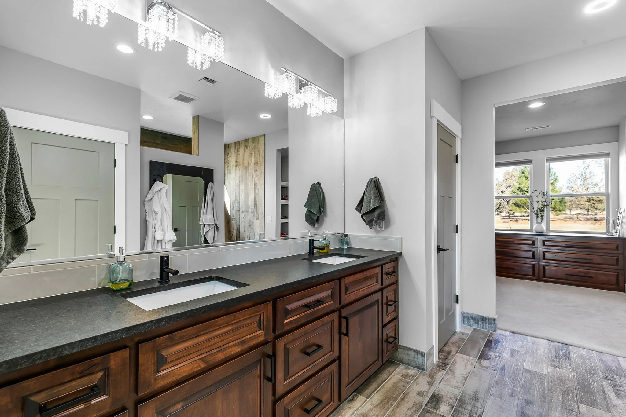 Modern bathroom with double vanity, dark wooden cabinets, black countertop, large mirror, white double sinks, chrome fixtures, wall-mounted light fixtures, towels hanging, and a large window overlooking a landscape.