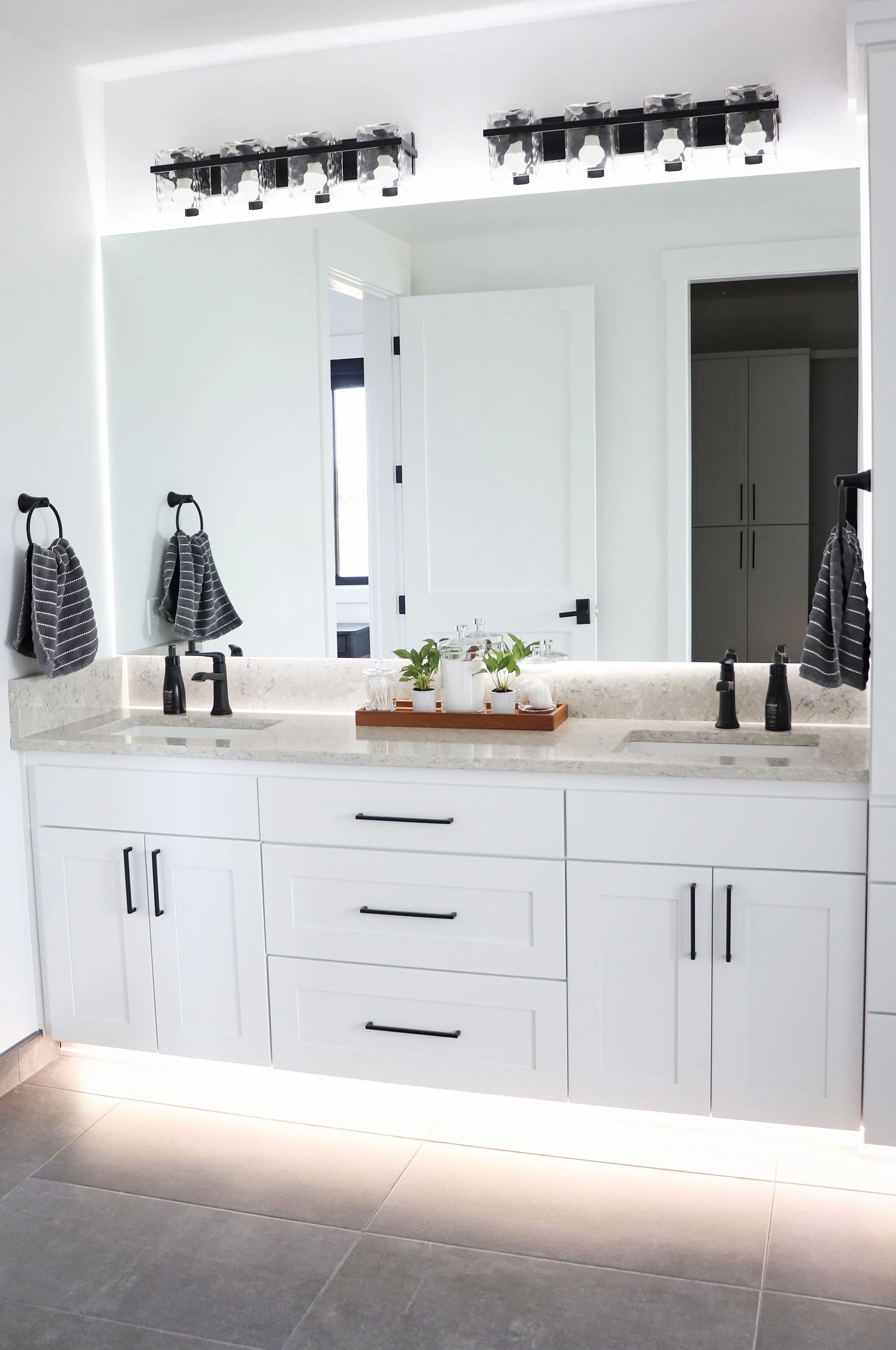 Modern bathroom with white vanity, black hardware, two sinks, large mirror, towels, plants, and overhead lighting.