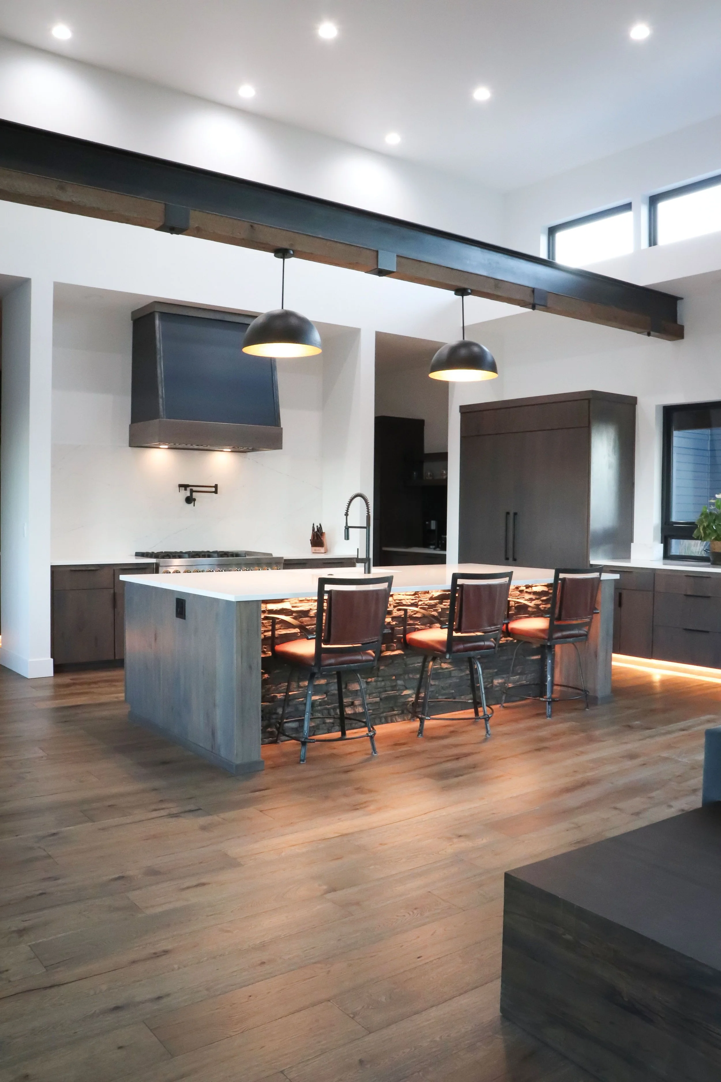 Modern kitchen with a large island, four bar stools, black pendant lights, and wooden flooring.