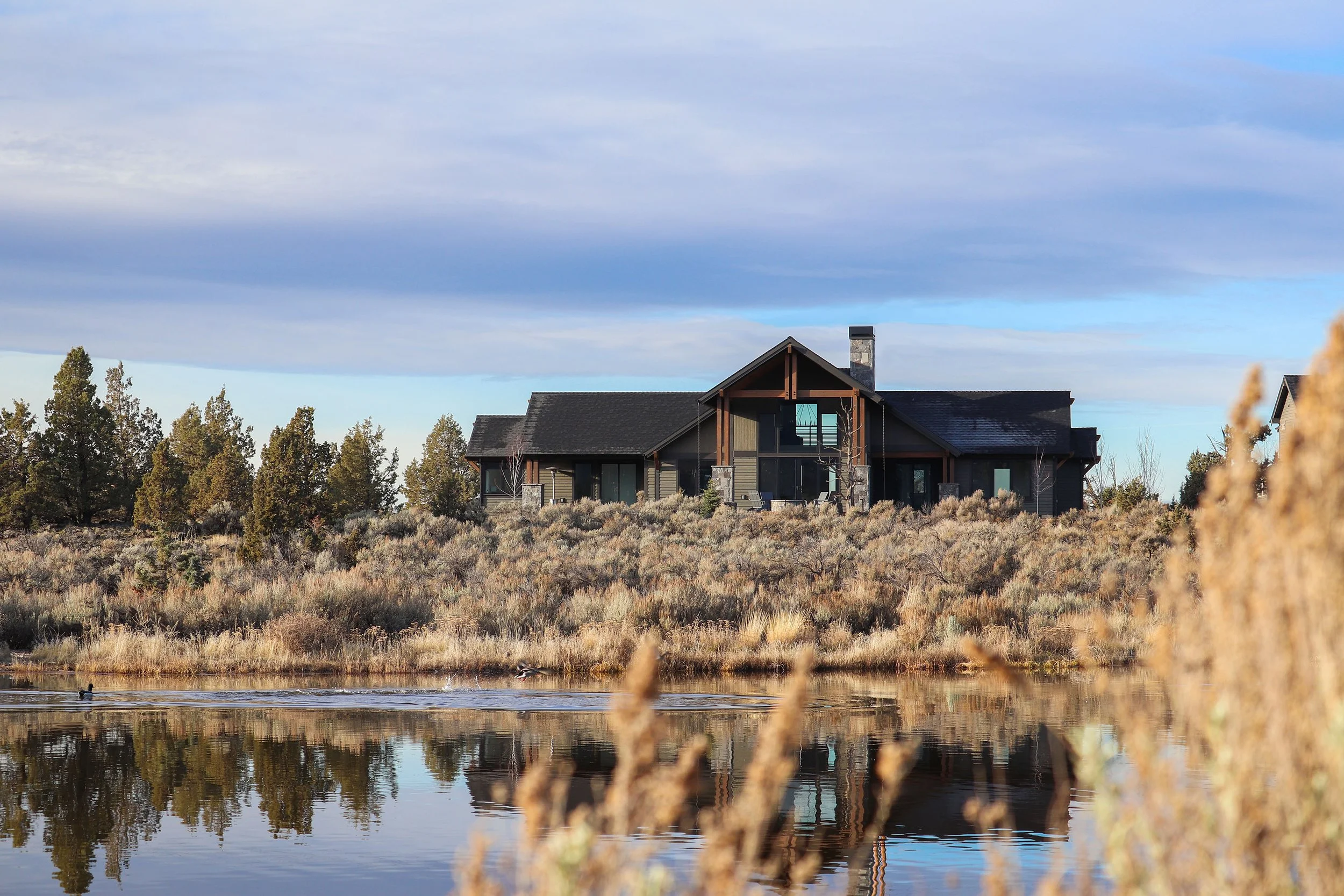 A large modern house with dark exterior walls and a stone chimney situated on a hill, with a pond in the foreground reflecting the house and surrounding shrubs and trees under a partly cloudy sky.
