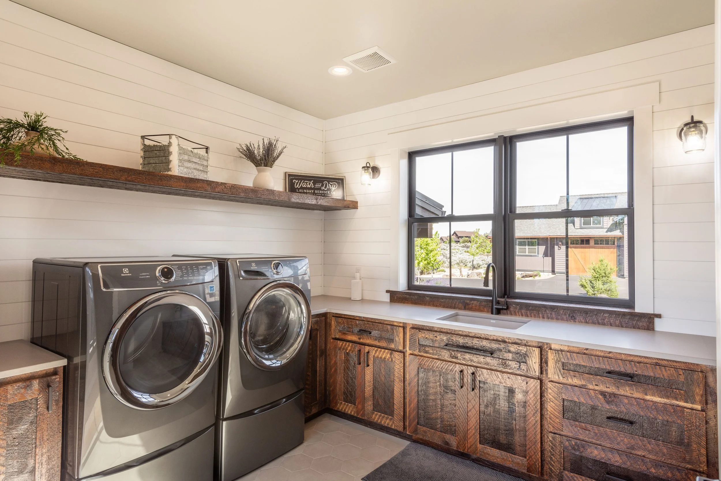 A laundry room with two front-loading washing machines, wooden cabinets, a rectangular sink under a large window, and decorative items on a wooden shelf.