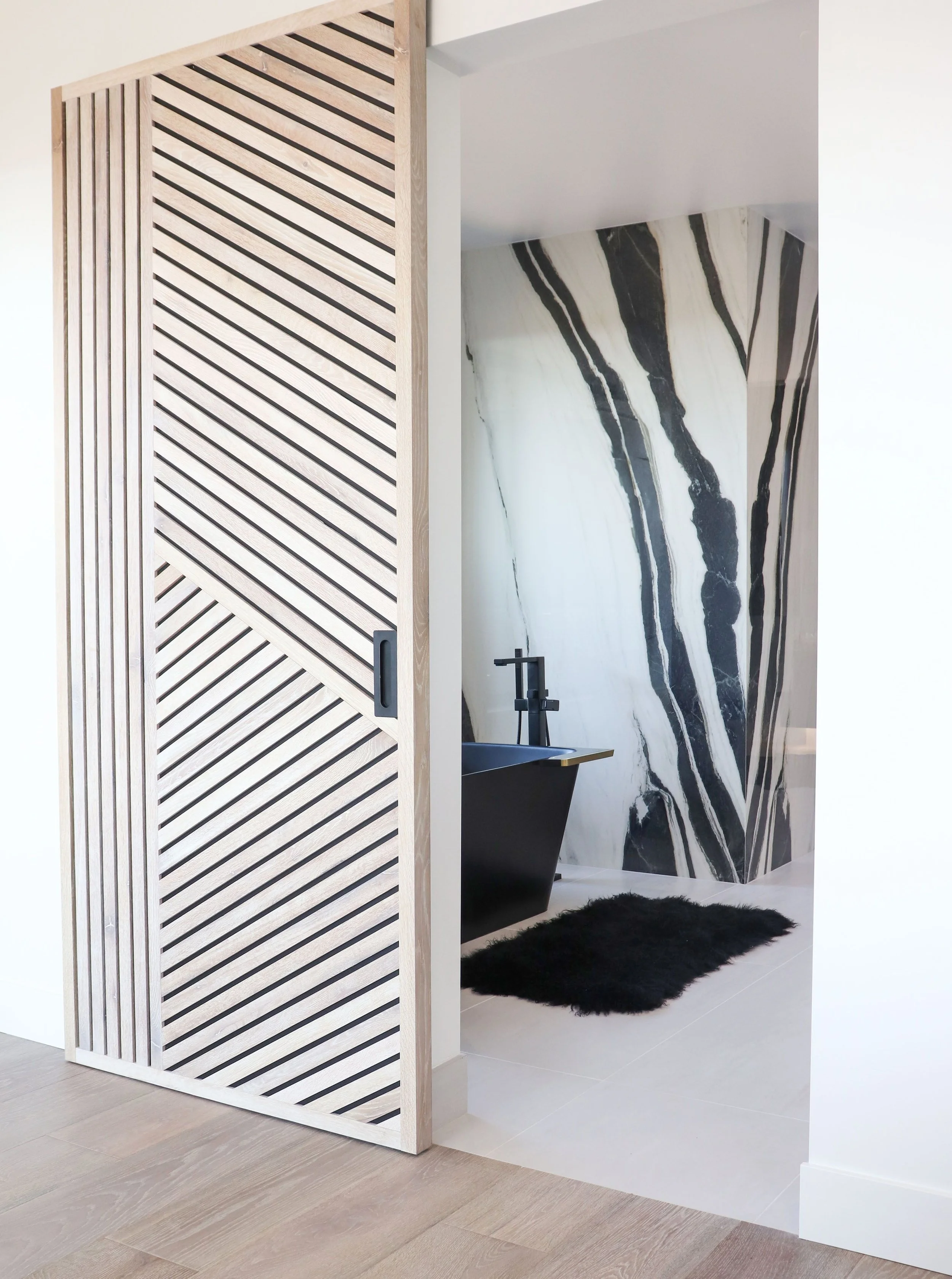 A modern bathroom with a black freestanding bathtub, black faucet, black fluffy rug, black and white marbled wall, and a sliding door with diagonal striped wood design.