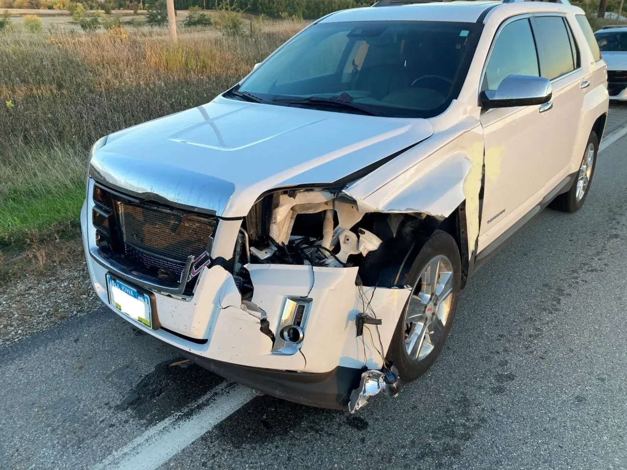 Damaged silver SUV with front-end impact, missing grille, exposed internal components, and a broken headlight on the driver's side, parked on the side of the road.
