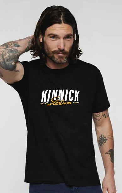Man with shoulder-length brown hair and tattoos wearing a black T-shirt with 'KUNICK Stadium' logo, posing with his right arm behind his head.