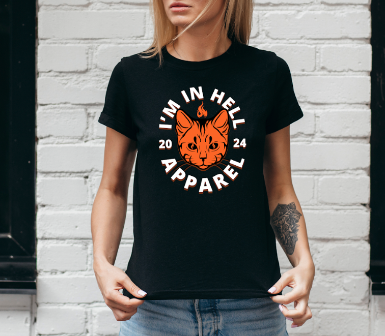 A woman wearing a black T-shirt with a graphic of an orange cat's face and the text "I'm in hell apparel" along with the year 2024. She is standing against a white brick wall.