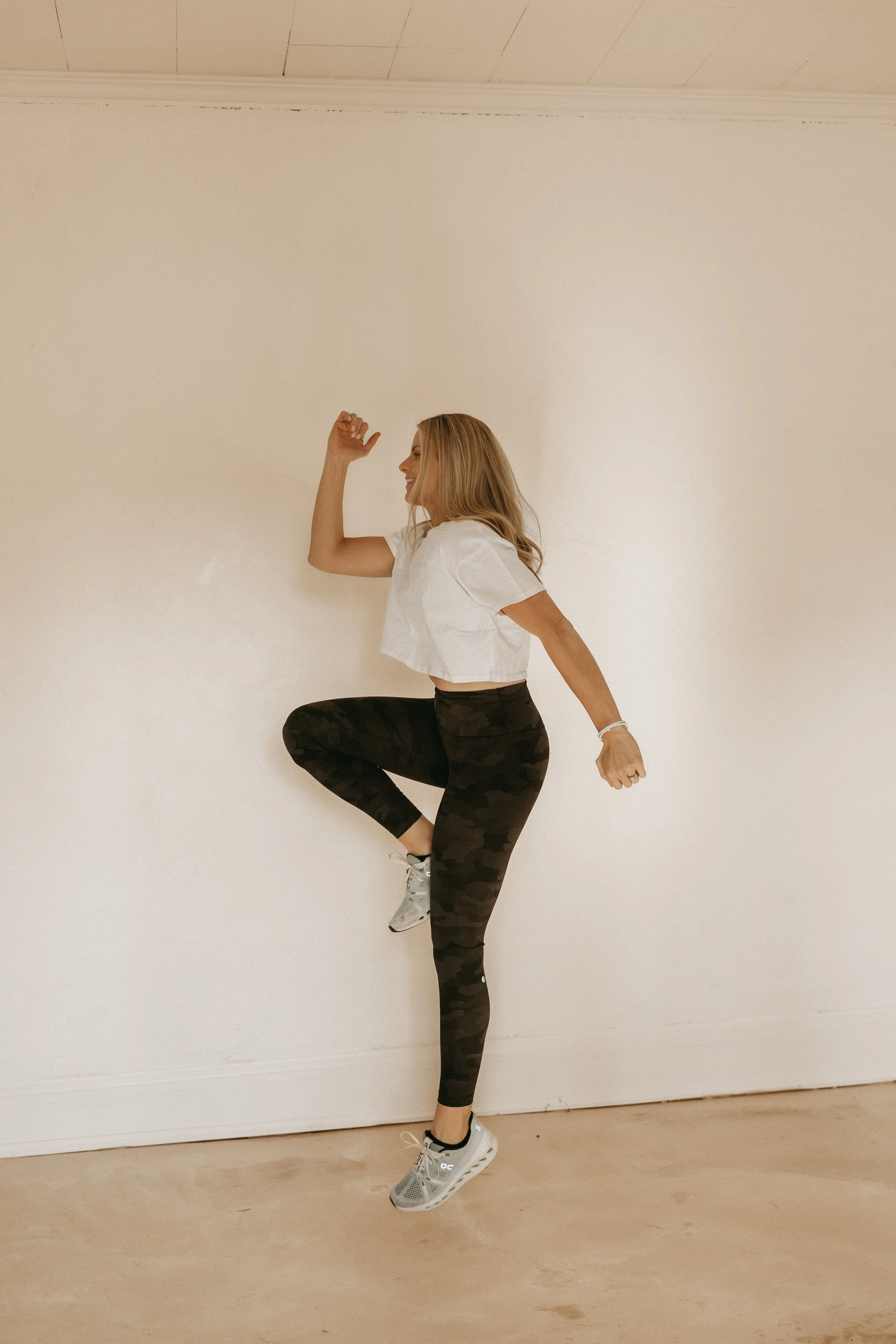 A woman in a white crop top and black camo leggings is performing a yoga pose against a plain wall, balancing on one leg with the other raised and bent at the knee.