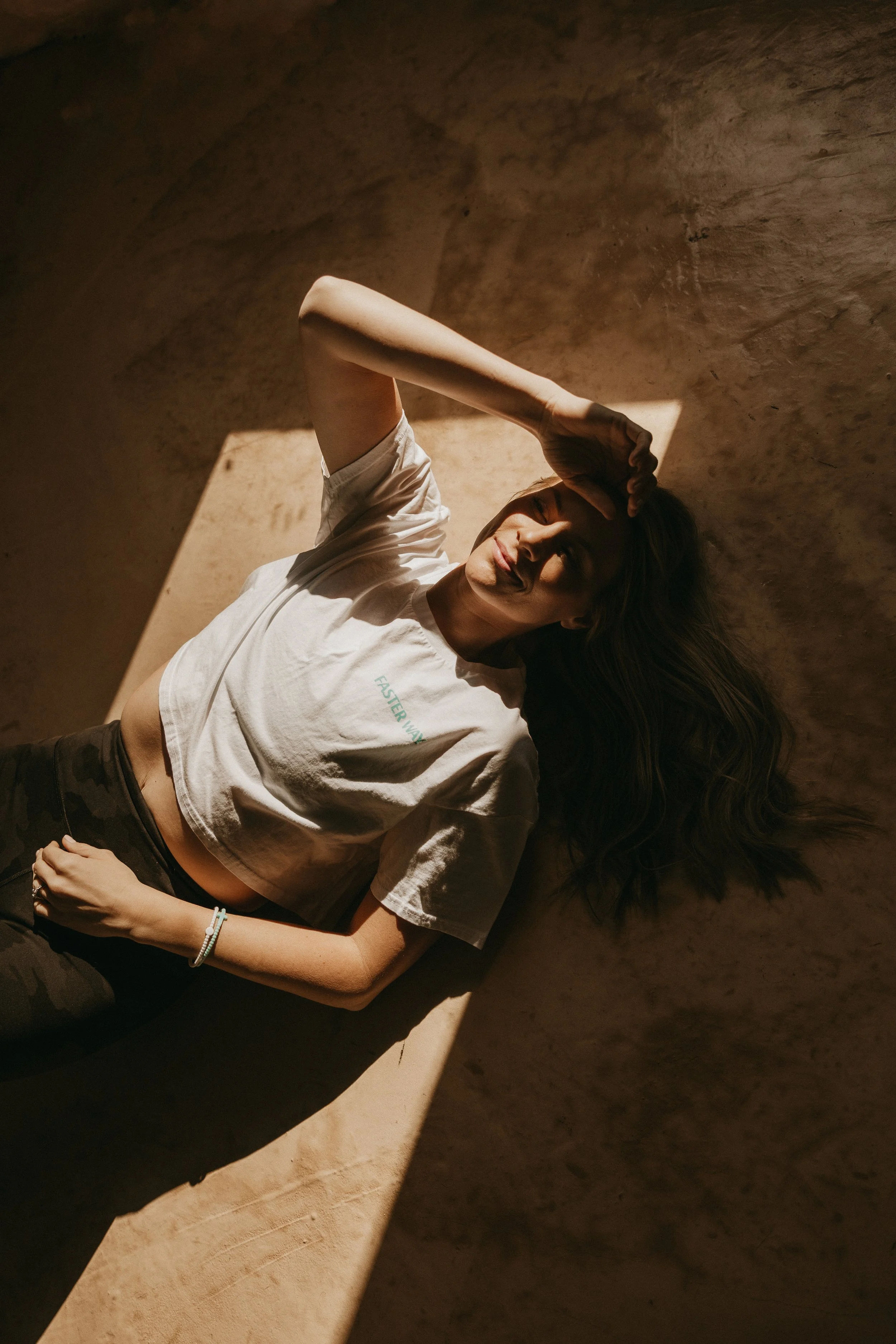 A woman lying on a sandy surface, with her eyes closed and hand on her forehead. She is wearing a white t-shirt and dark pants, with her long hair spread out on the ground. Sunlight casts shadows across her face and body.