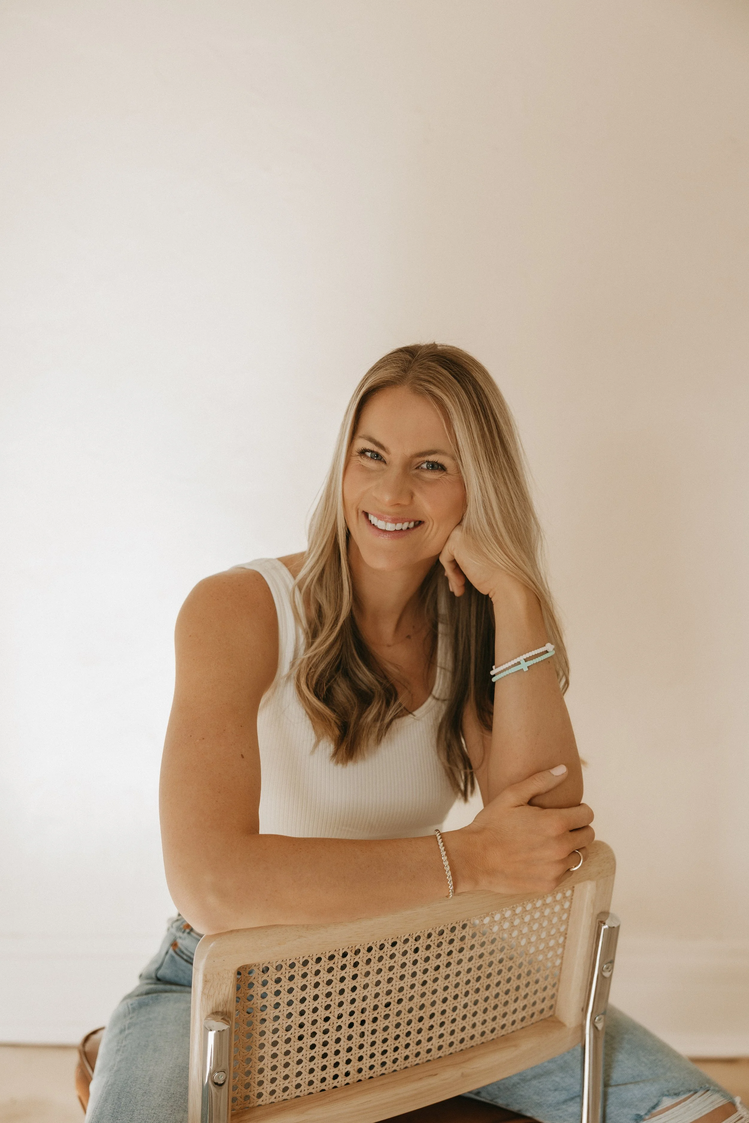 Founder of femwell, Allie Janszen, wearing a white tank top, seated on a beige chair with a woven back, against a plain off-white wall.