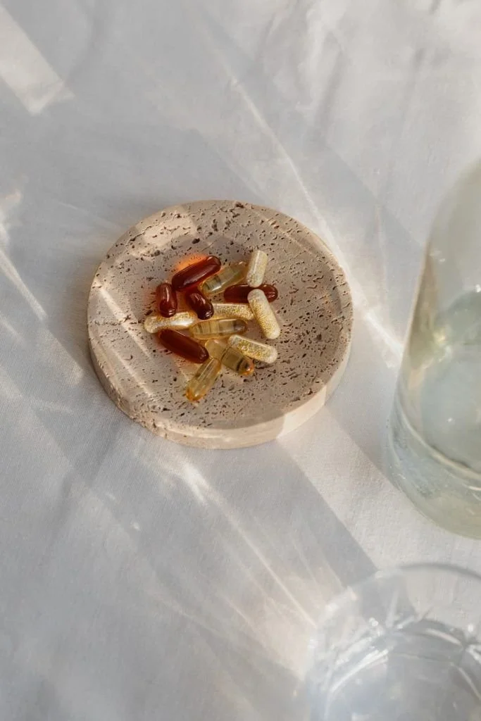 A small round stone or tile with various capsules and pills on top, positioned on a white fabric surface beside a glass of water.