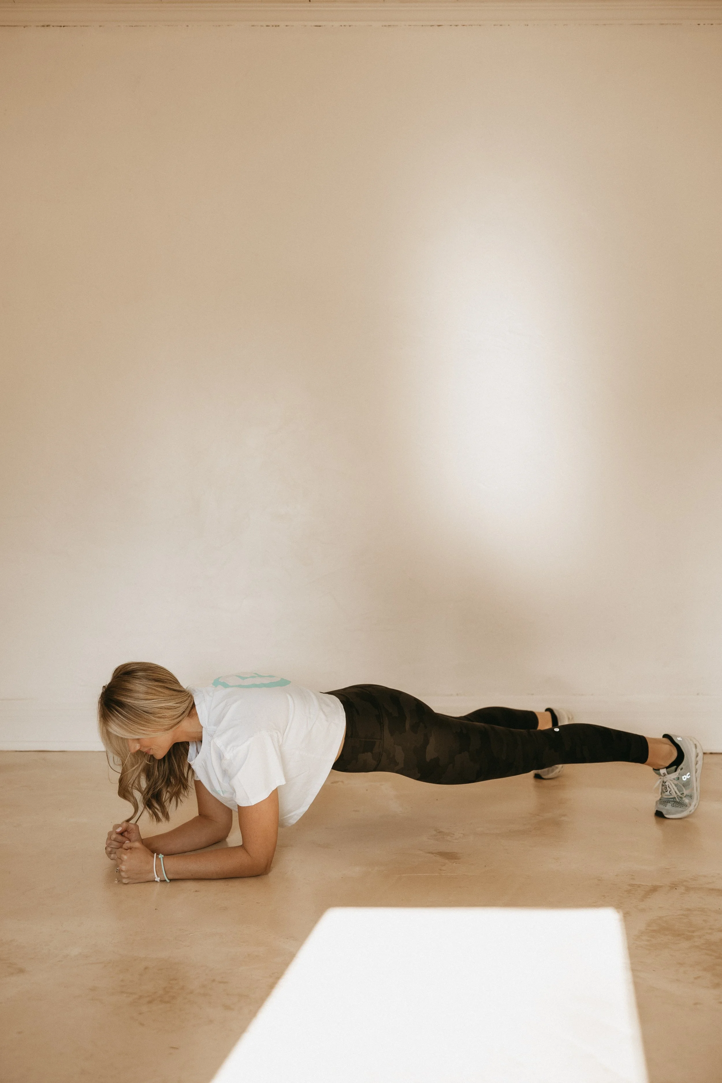 A woman with long blonde hair performing a plank exercise on the floor in a gym or studio. She is wearing a white t-shirt, black camouflage leggings, and sneakers.