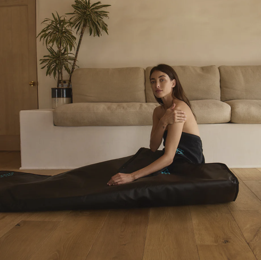 A woman sitting on a black padded object on a wooden floor, with a beige sofa and a potted plant in the background.