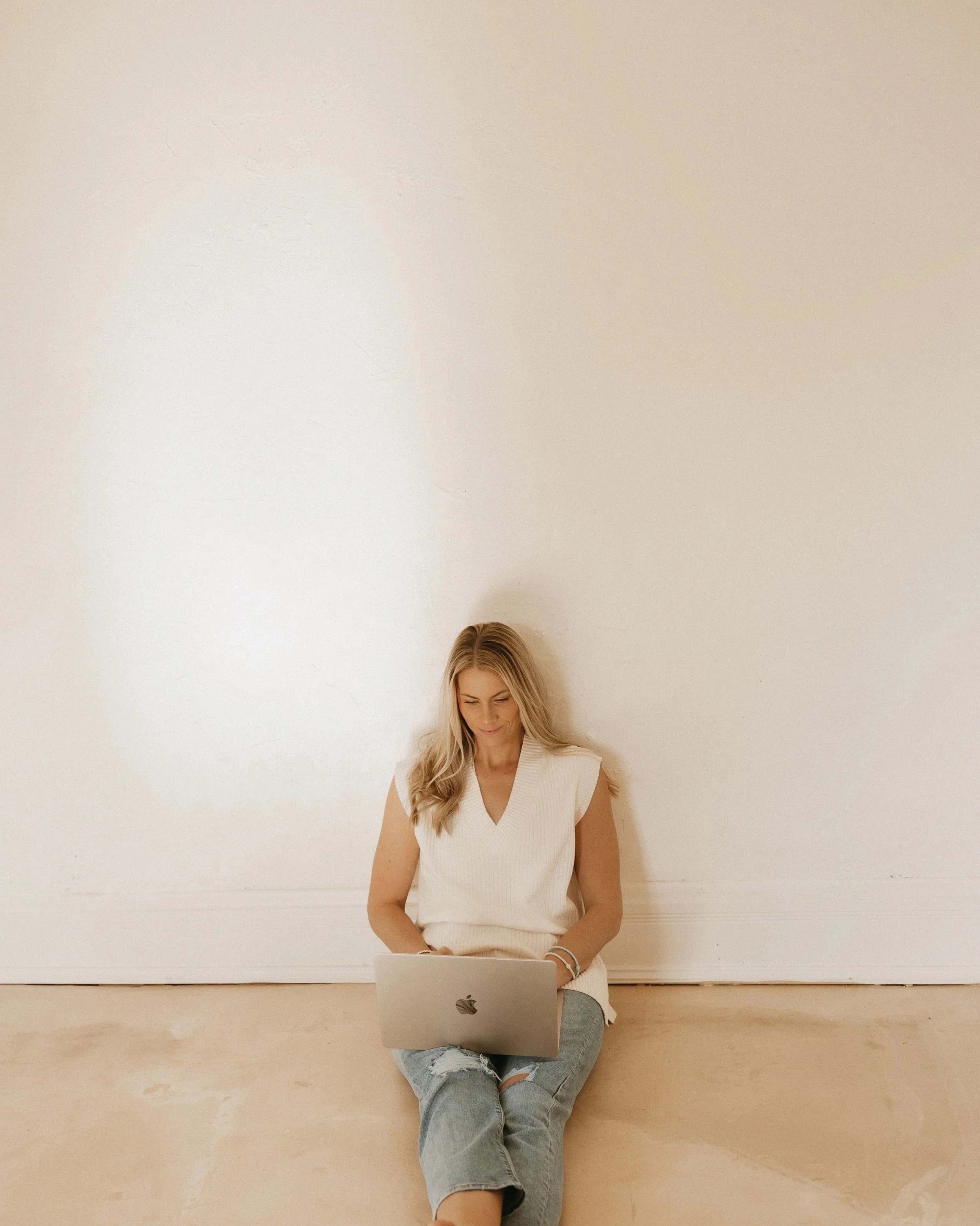 Founder of femwell, Allie Janszen, in a sleeveless beige top and ripped jeans sitting on a ledge, using a silver laptop in front of a plain, off-white wall.