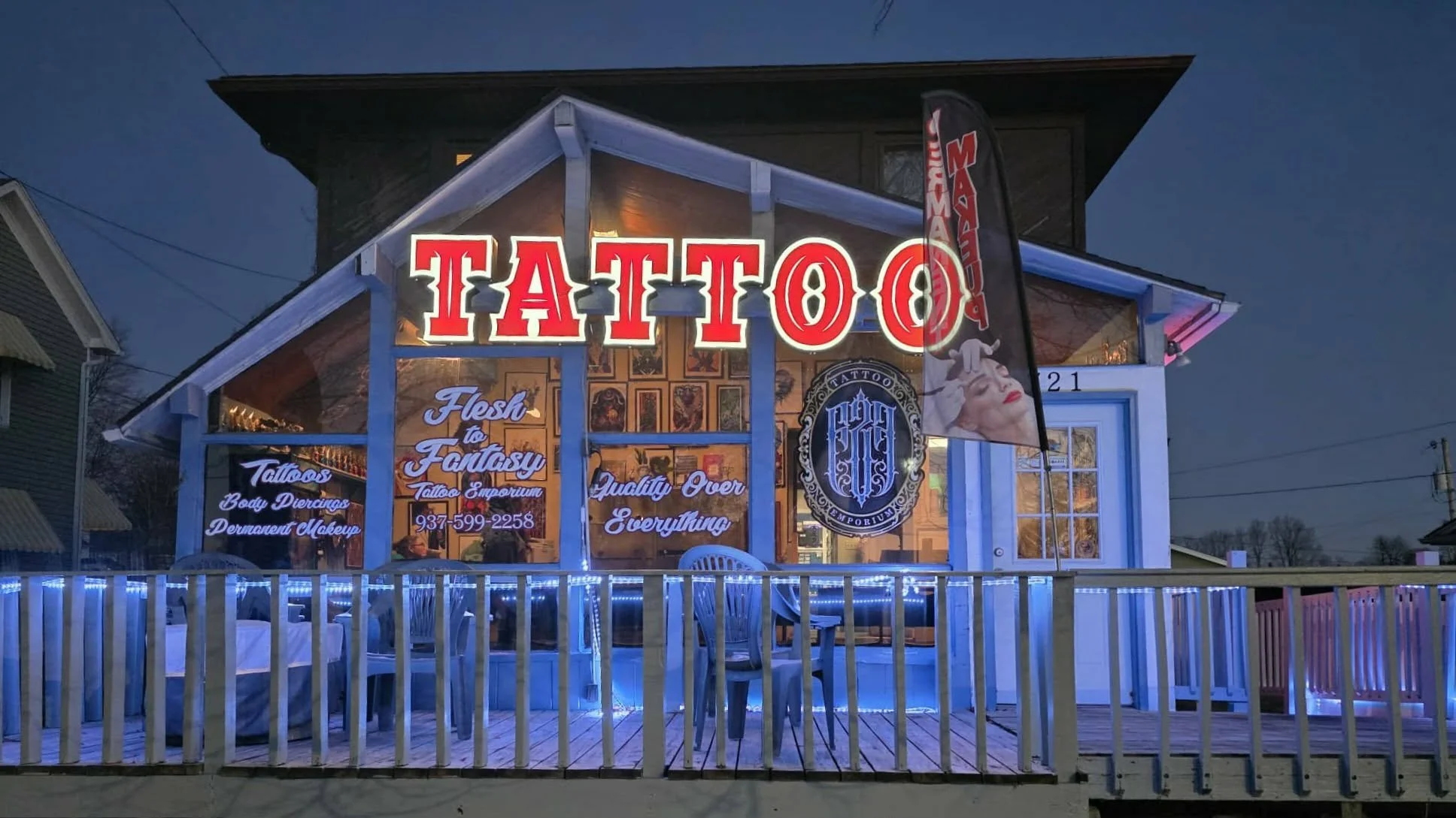 A tattoo shop with large illuminated red and white sign that reads 'TATTOO' in front of a building at dusk. The shop has large windows displaying various art tattoos and neon lights; a black banner with the shop's logo and name, and a white porch with chairs and a railing decorated with string lights.