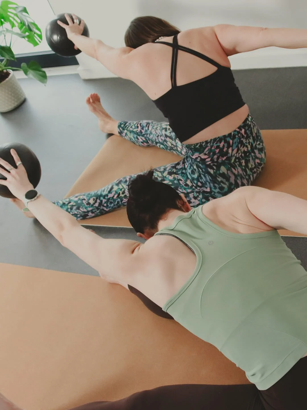 Two women doing pilates exercises on mats with medicine balls in a bright room with a window and a potted plant.