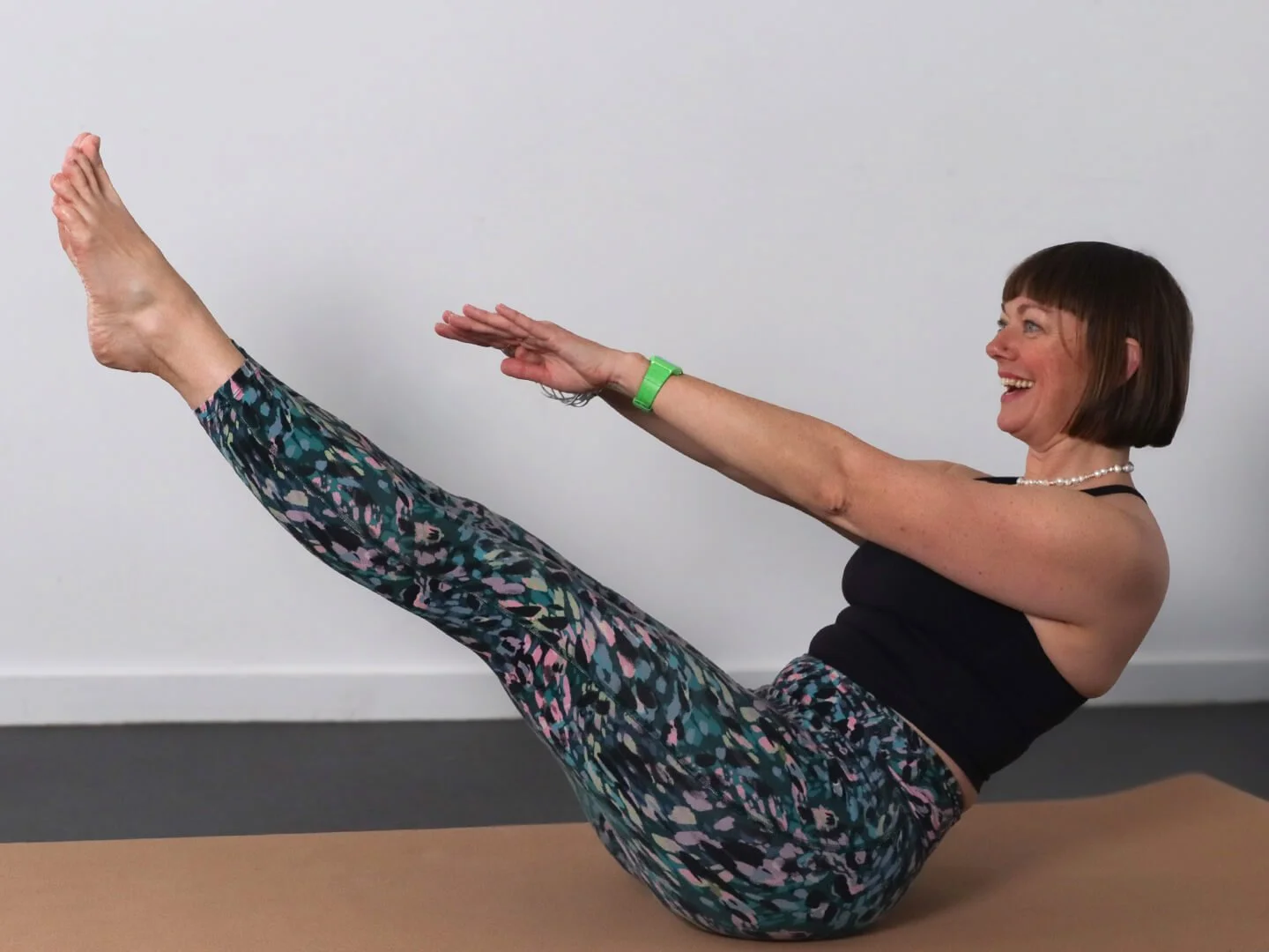 Woman doing a boat pose yoga stretch, lying on her back with arms extended forward and legs elevated, against a plain wall background.