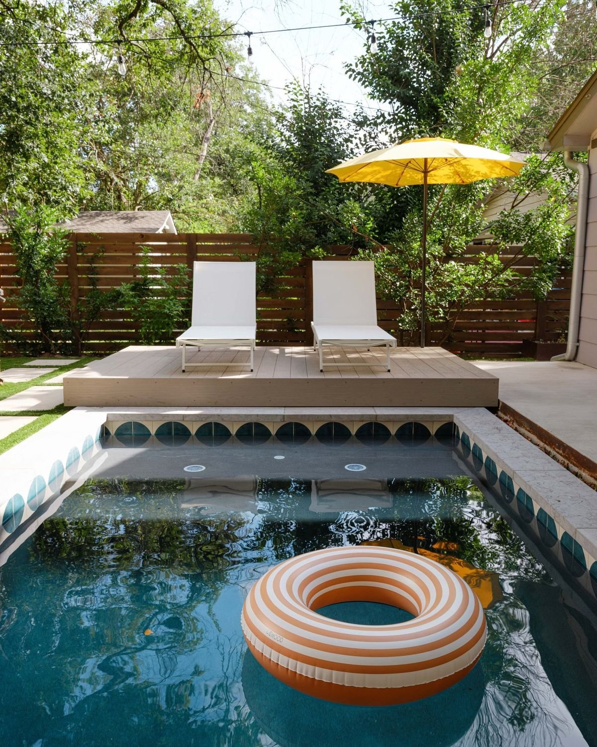 Private backyard plunge pool with blue tiled accents, a raised wooden sun deck with two white lounge chairs, and a yellow umbrella.