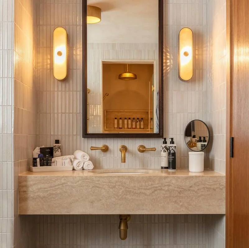 Luxury bathroom vanity made of light-colored stone with brass fixtures, vertical white tile, and modern pill-shaped light sconces.