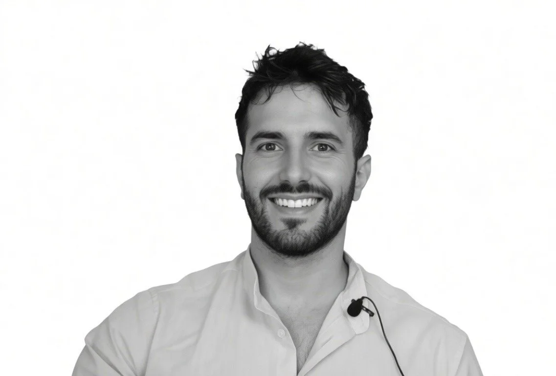 Un uomo sorridente con barba e capelli castani corti, indossa una camicia bianca e ha un microfono appoggiato sulla camicia.