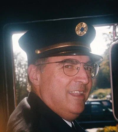 A man in a Fire Cheif officer uniform with a cap and glasses smiling inside a vehicle.