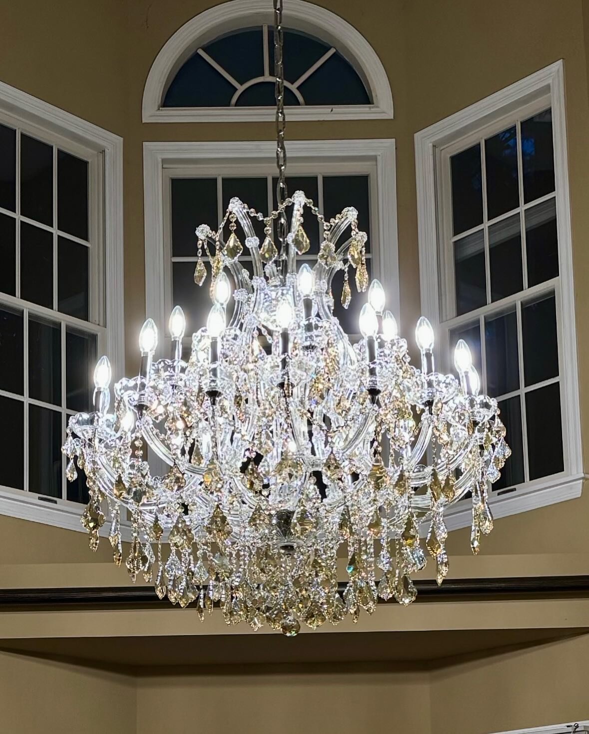 A large, ornate chandelier with numerous crystal ornaments and candle-shaped light bulbs hanging from a ceiling in a room with large windows and beige walls.