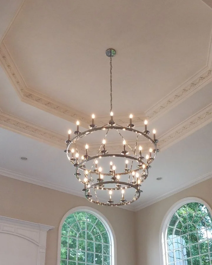 Large chandelier with multiple rings of small lights hanging from a ceiling with crown molding in a bright room with arched windows.