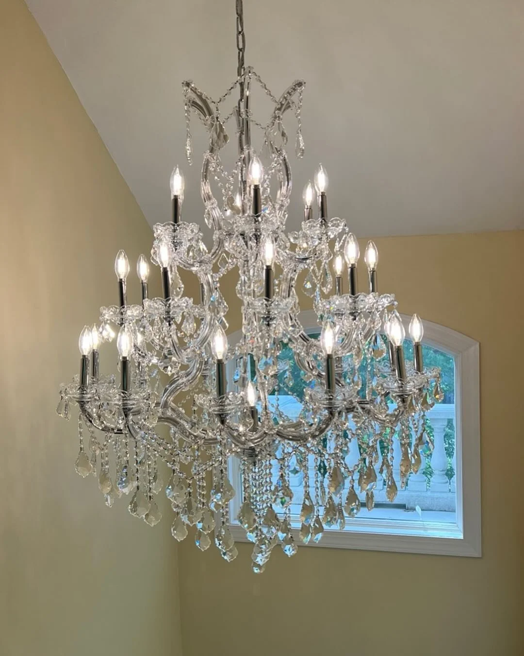 A large, ornate crystal chandelier with multiple arms and candle-shaped lights hanging from a ceiling, next to a window with a view of a porch.