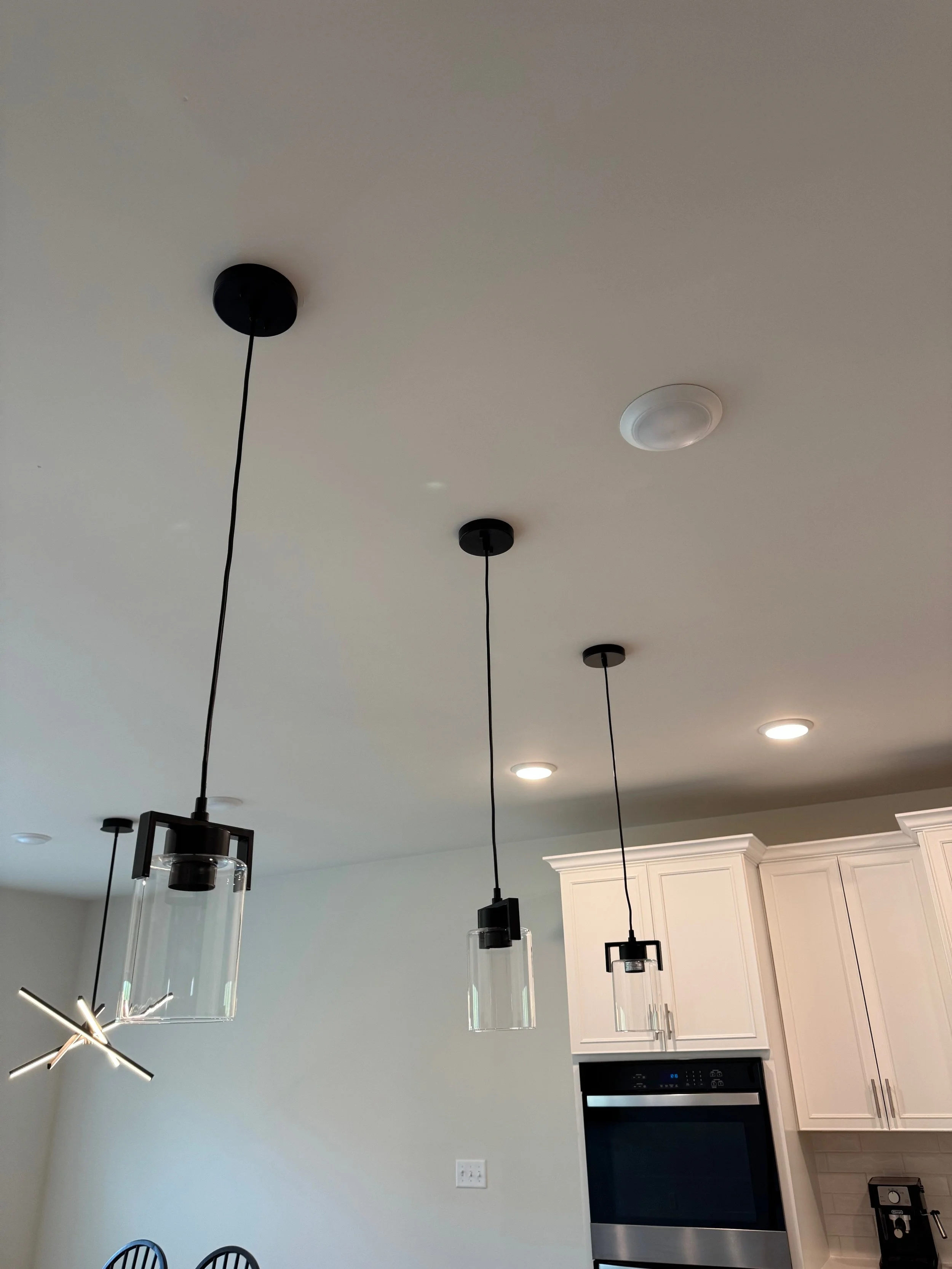 Modern kitchen ceiling with three black pendant lights with glass shades, and a white ceiling-mounted smoke detector; part of white kitchen cabinets and stainless steel oven are visible.