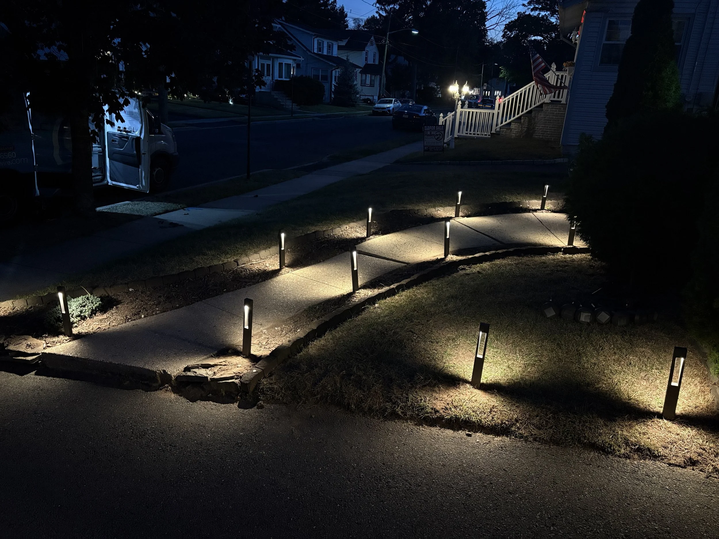 Nighttime view of a sidewalk with solar-powered pathway lights illuminating the concrete and grass, with houses and cars visible in the background.